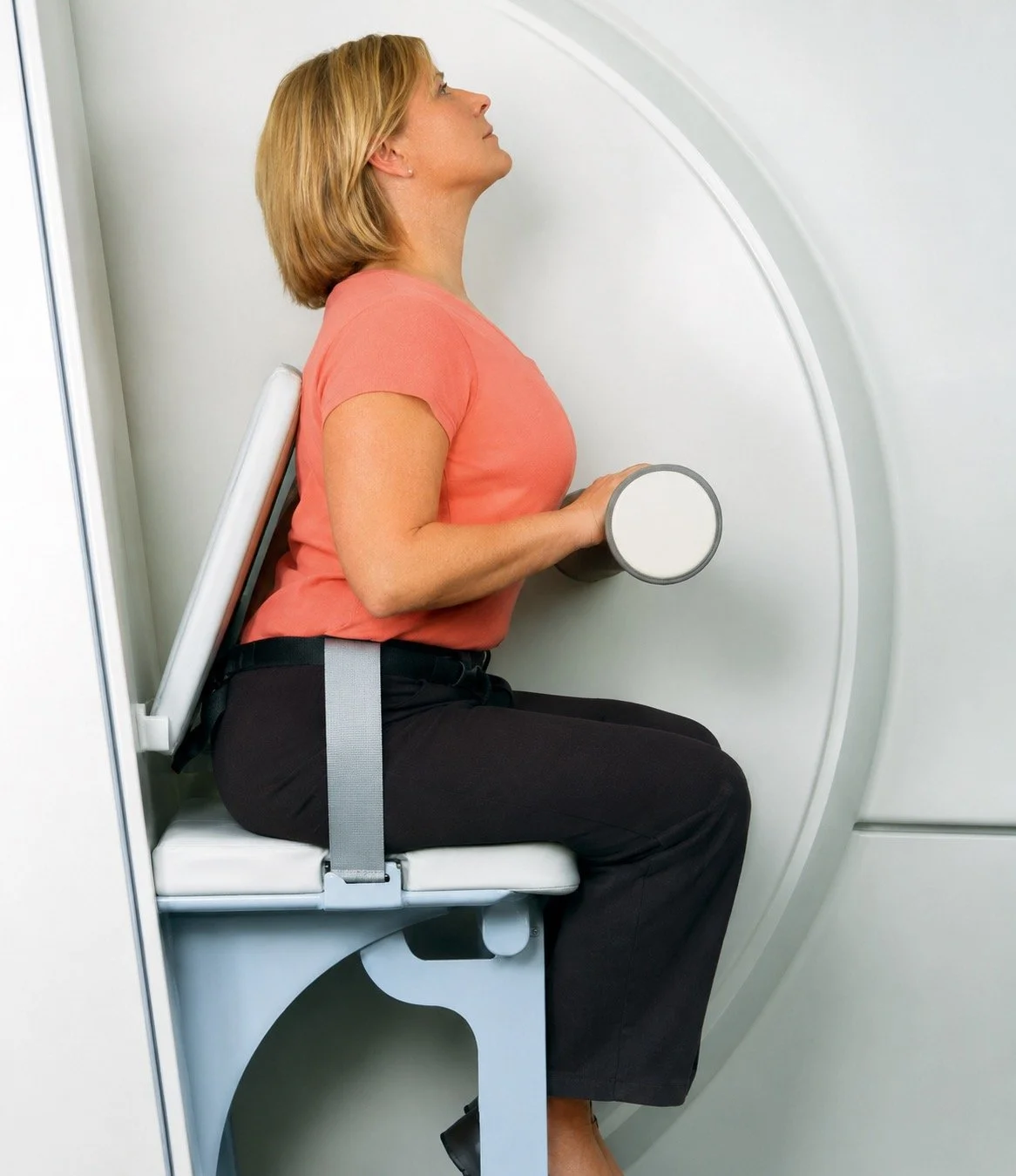 A woman undergoing a medical imaging procedure, sitting on a chair with a device behind her back and holding a dumbbell.
