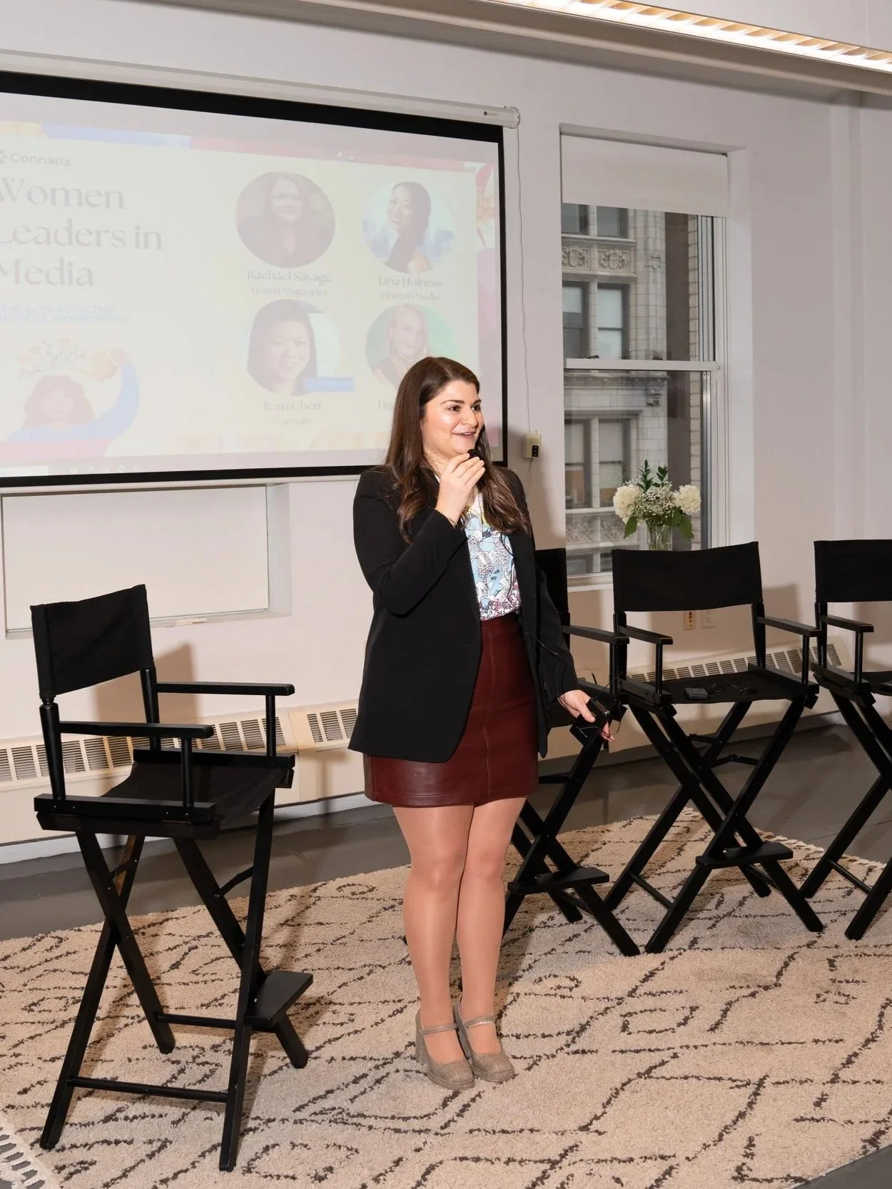 A woman stands in front of a presentation screen in a conference room, speaking or preparing to speak, with chairs arranged for an audience, window with a view of buildings outside, and a vase with flowers on the window sill.