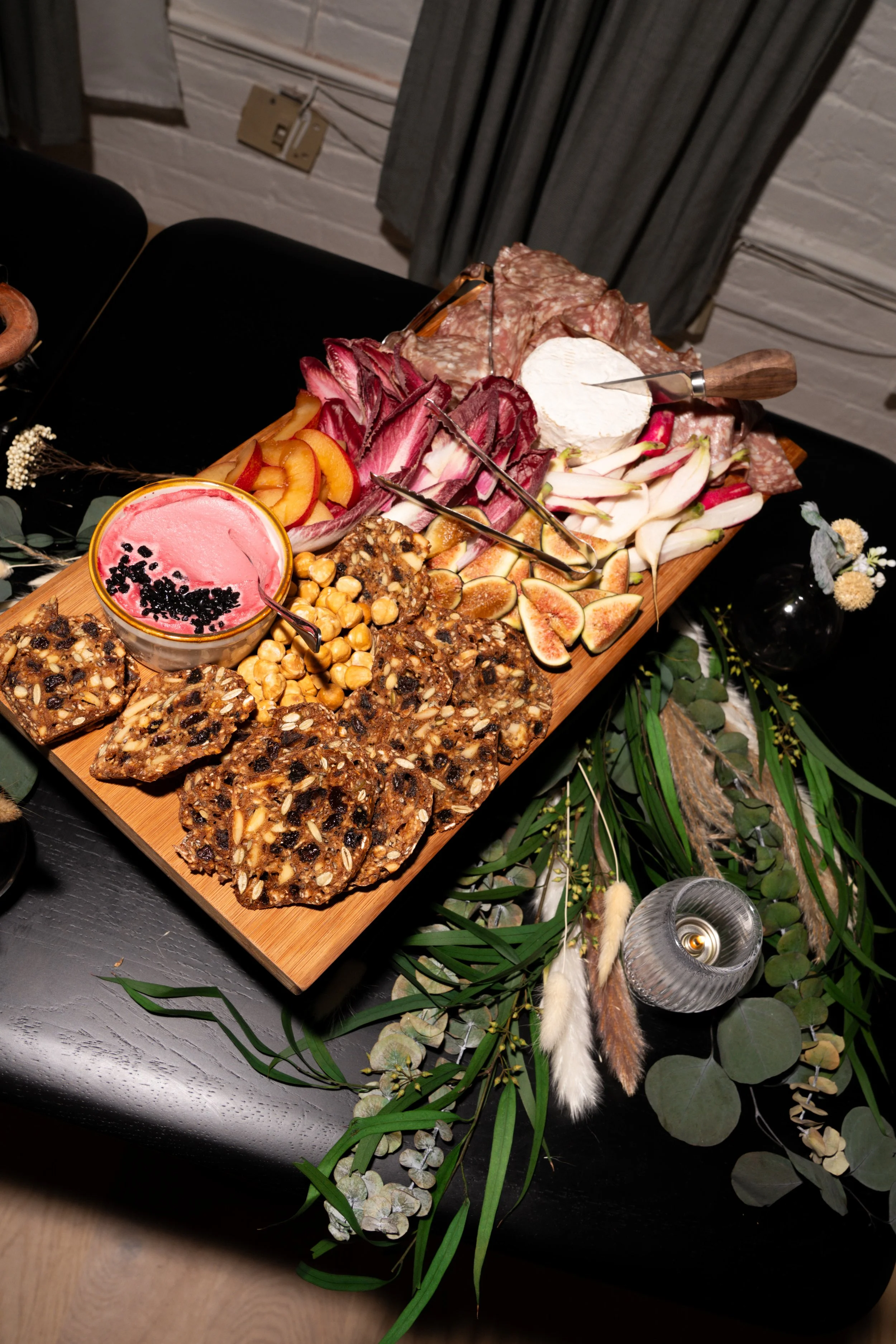 A charcuterie board with various sliced meats, cheeses, fruits, crackers, and spreads, placed on a black table decorated with greenery and floral arrangements.