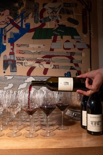 A hand pouring red wine into a row of wine glasses on a wooden table in front of an abstract painted wall.