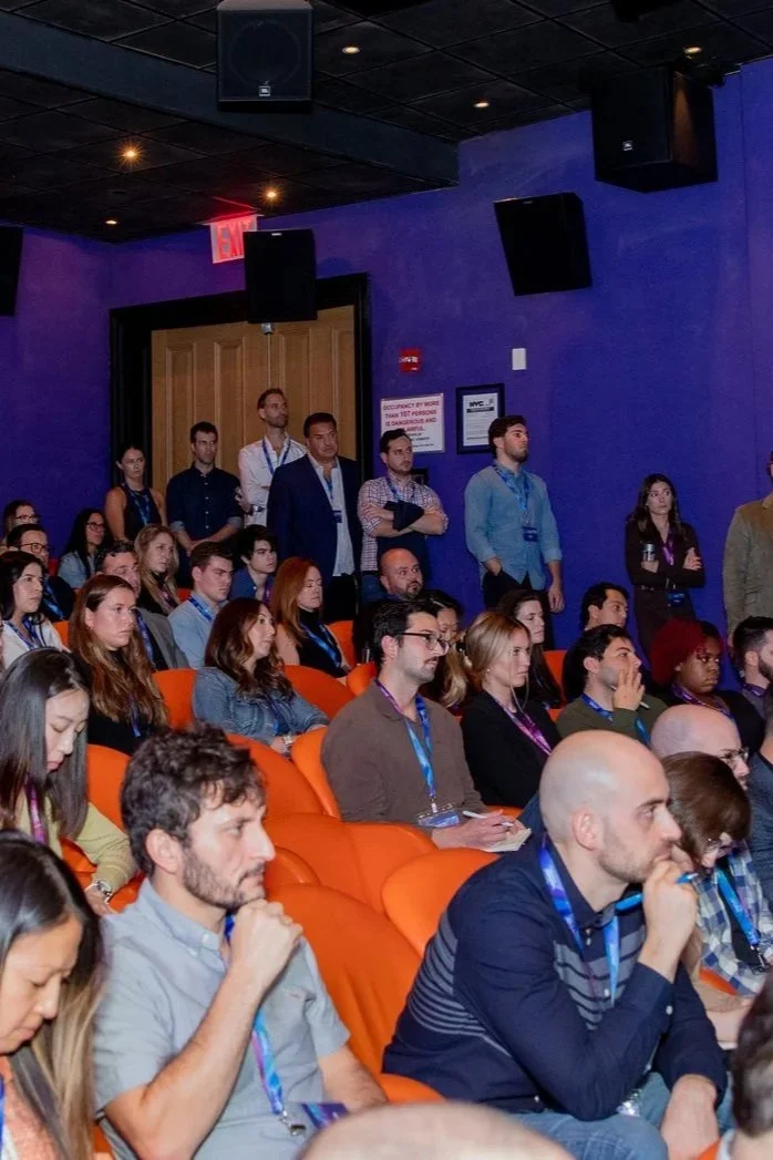 Audience at a conference or seminar seated in orange chairs, listening attentively, with some standing near the back of the room, in a blue-walled auditorium.