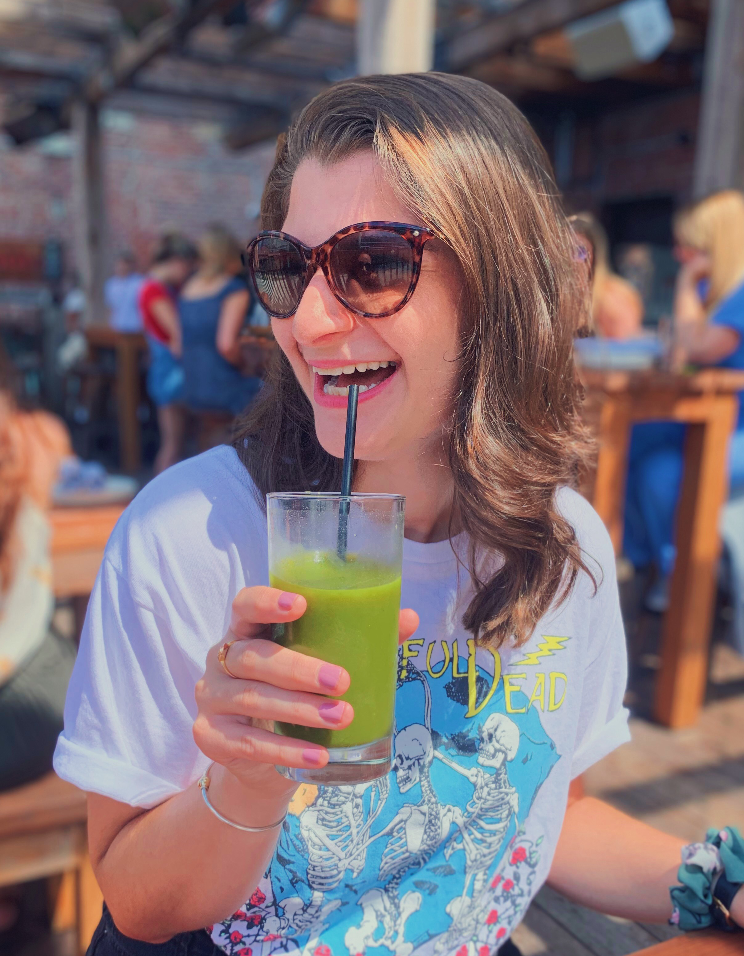 A woman with long brown hair wearing large tortoiseshell sunglasses and a white graphic t-shirt smiling and drinking a bright green beverage with a straw at an outdoor setting, with other people in the background.