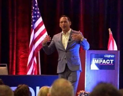 Man in gray suit speaking at a conference podium with an American flag in the background and a Cigna banner to the side.