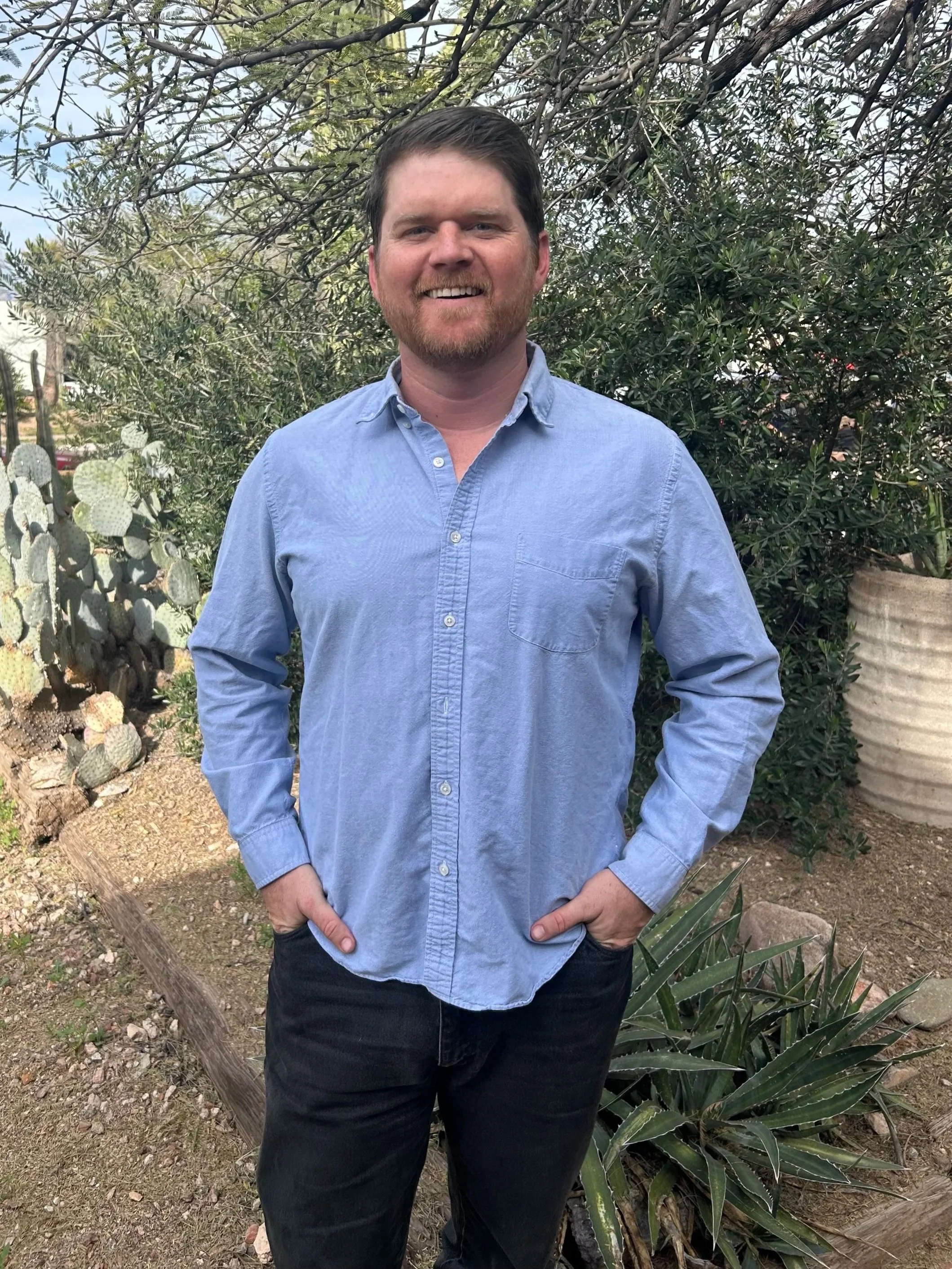A man with dark hair and a beard smiling, wearing a light blue button-up shirt with rolled-up sleeves and black pants, standing outdoors in front of a bush with green leaves and a large cactus, with a wooden log on the ground and a large plant with long pointed leaves in the foreground.