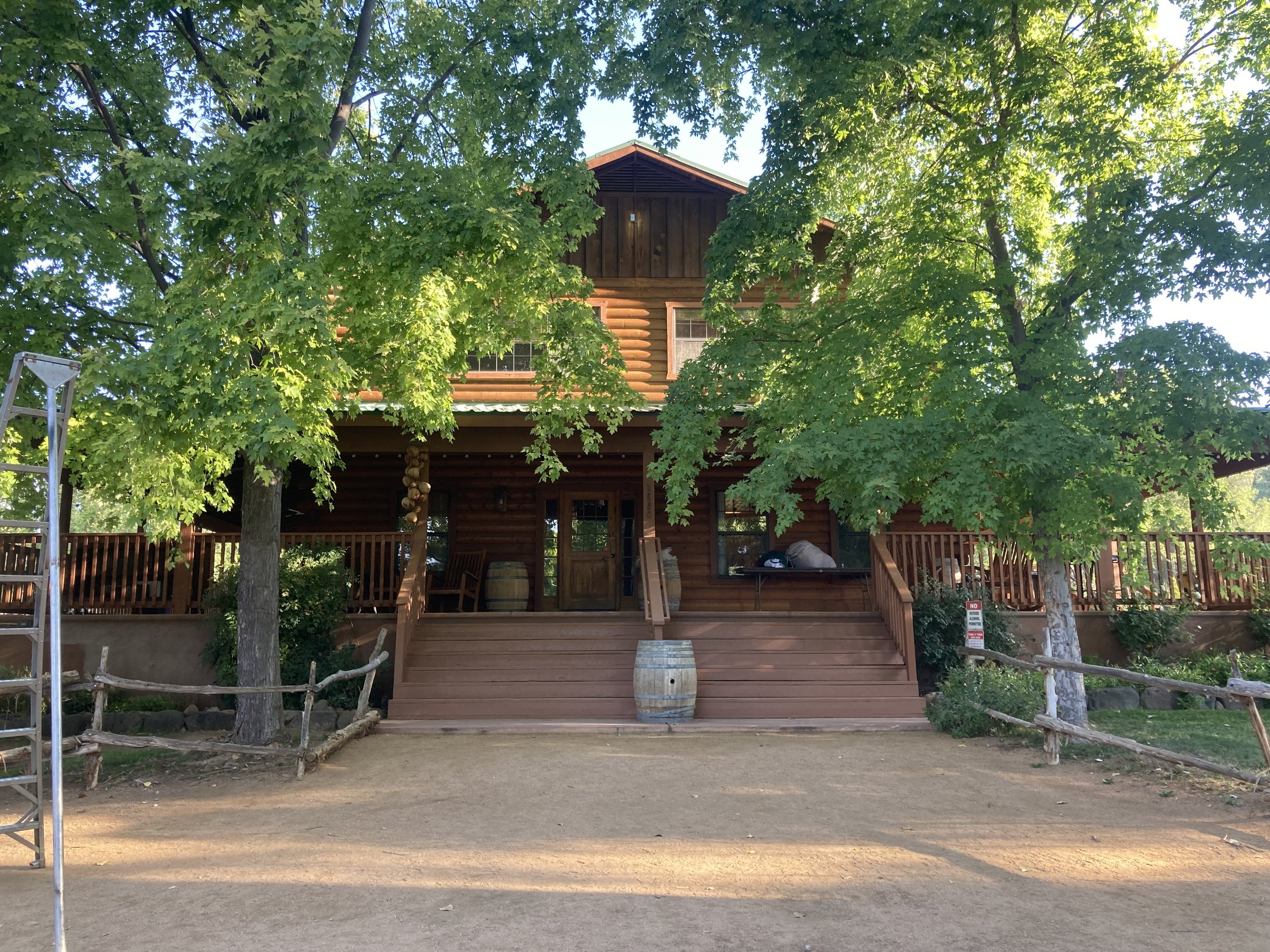 A two-story log cabin partially obscured by large green maple trees in front. The cabin has a front porch with steps, wooden deck, and barrels for decoration. A ladder is visible on the left side of the image, and a sign indicating no parking is on the right side. The scene appears to be in a wooded area with a natural dirt ground.