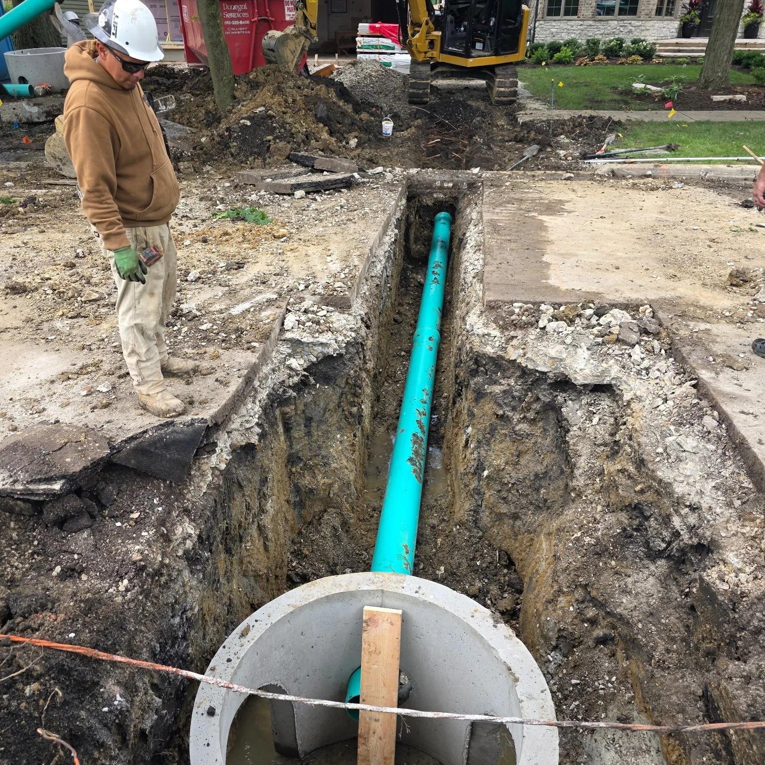 A construction worker wearing a white hard hat, sunglasses, a brown hoodie, and gloves stands next to a deep trench with a blue pipe inside. The trench is surrounded by soil and construction equipment, with a small excavator in the background.
