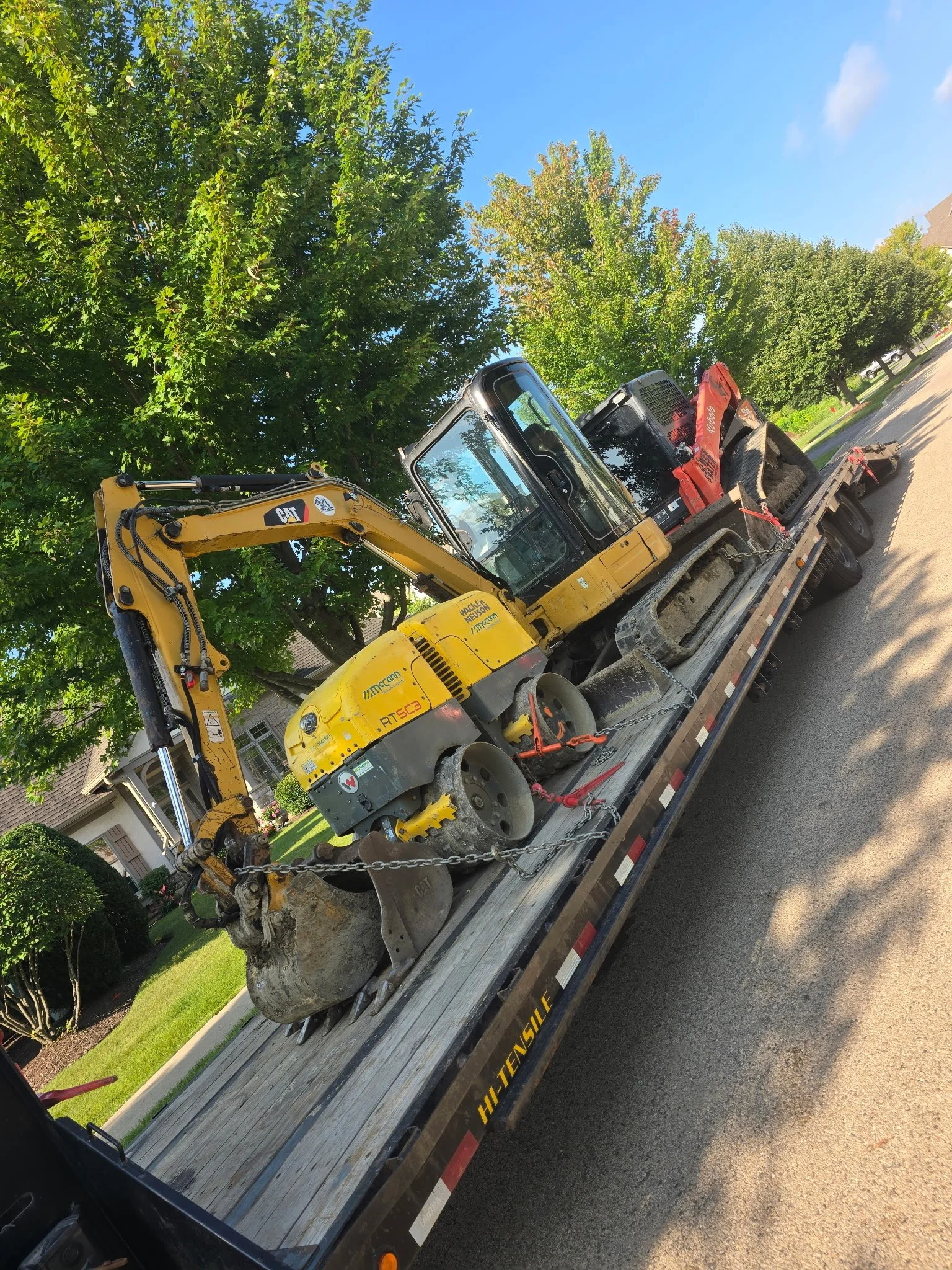A flatbed truck carrying construction equipment, including a yellow excavator, parked on a residential street lined with green trees and houses under a clear blue sky.