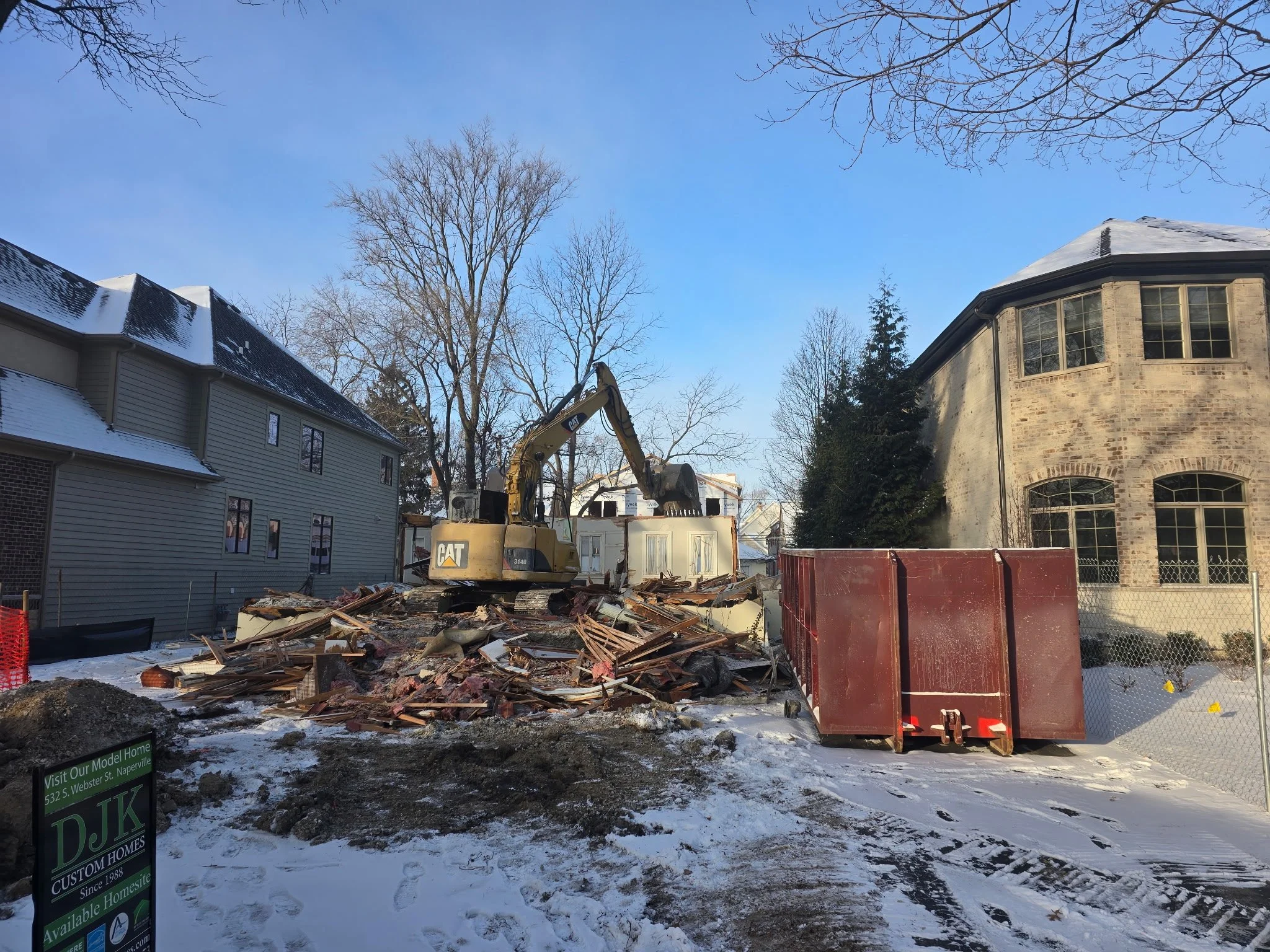 Construction site with a yellow excavator and debris, snow on the ground, neighboring houses 