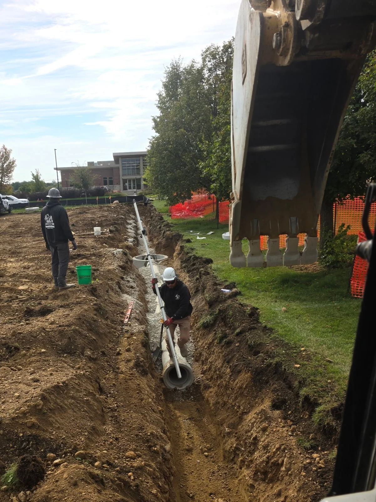 Construction workers installing a large pipe into a long trench at a construction site, with machinery and a building visible in the background.