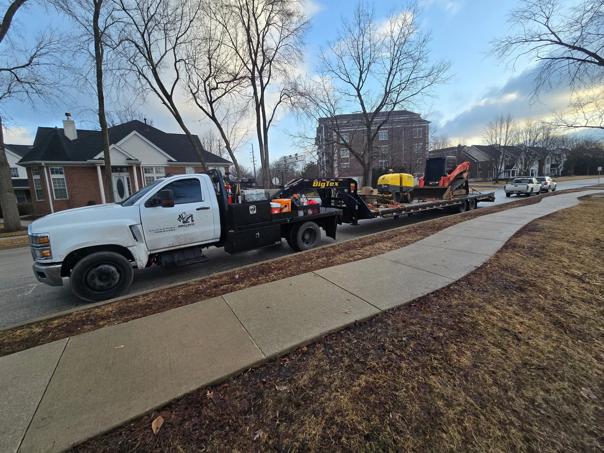A white flatbed truck parked on a residential street carrying construction equipment, including a small yellow excavator and a road roller. Houses and leafless trees are visible along the street.