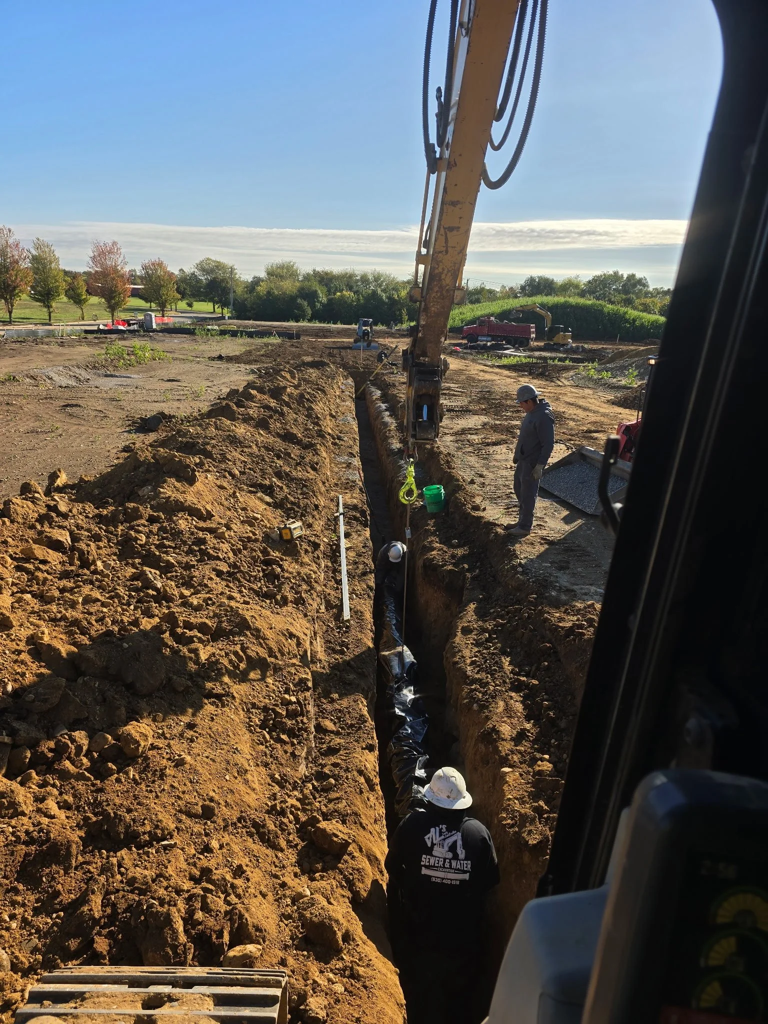 Construction workers at a trench, installing pipes underground, with excavator and trucks nearby, on a clear day with trees in the background.