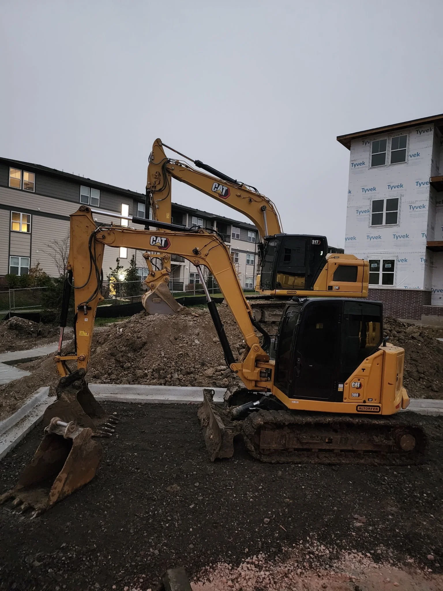 Construction site with two yellow excavators digging soil next to a building under construction, in an urban area during dusk.