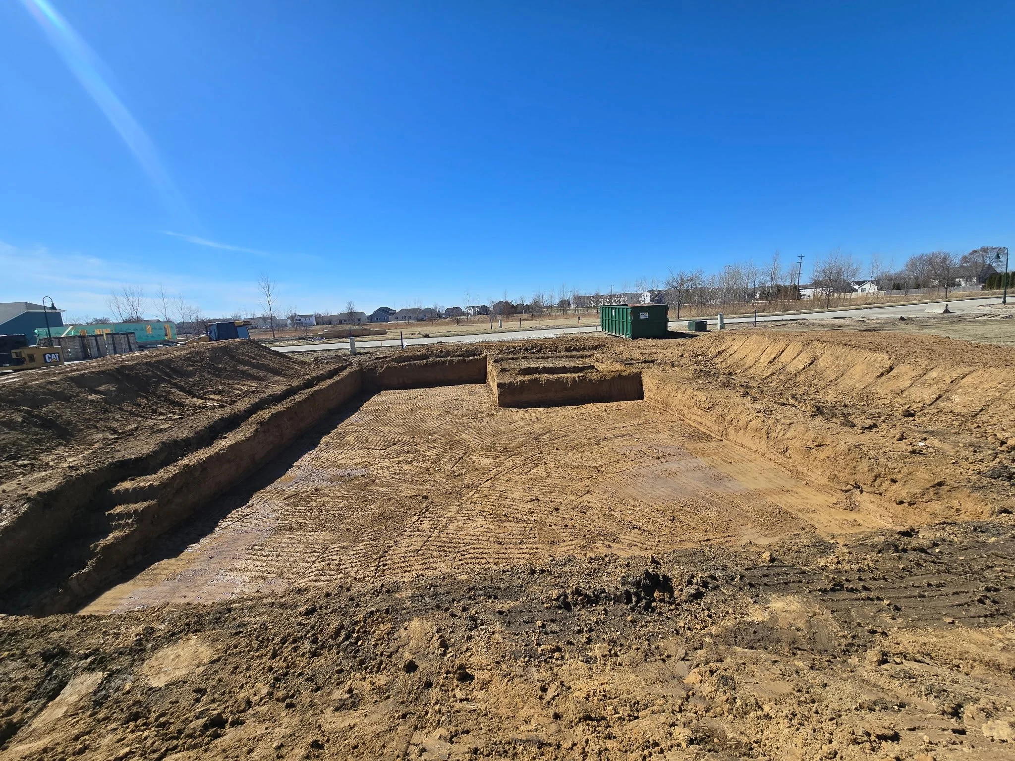 Construction site with excavated dirt and a green dumpster, with houses in the background on a clear, sunny day.