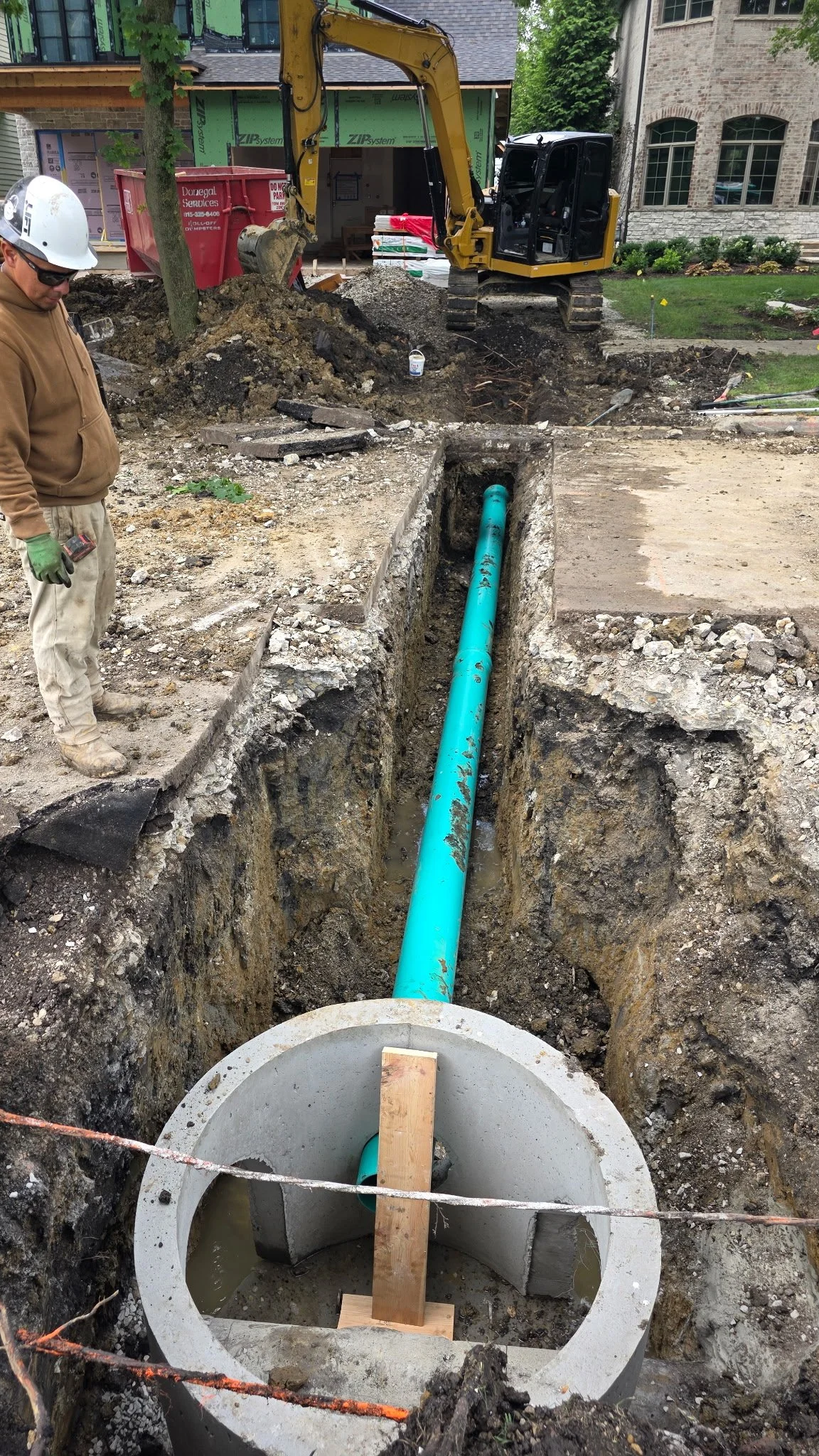 A construction site showing an open concrete sewer pipe with a blue pipe inside. A construction worker wearing a white helmet and brown hoodie stands nearby. In the background, an excavator is digging up the ground, and there are piles of dirt and construction materials.