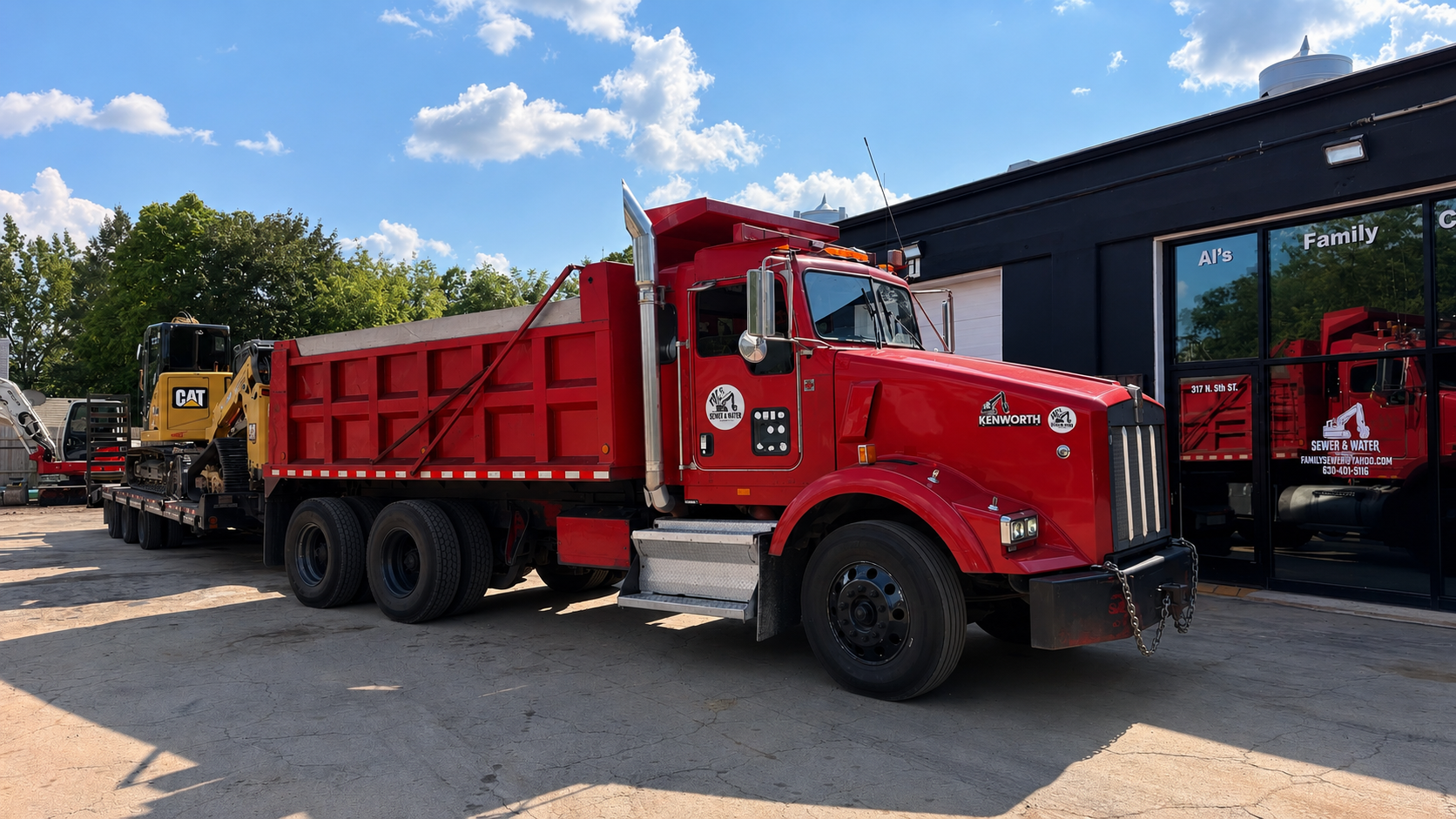 Red tow truck parked outside a building with large glass windows, alongside other construction equipment, under a partly cloudy blue sky.