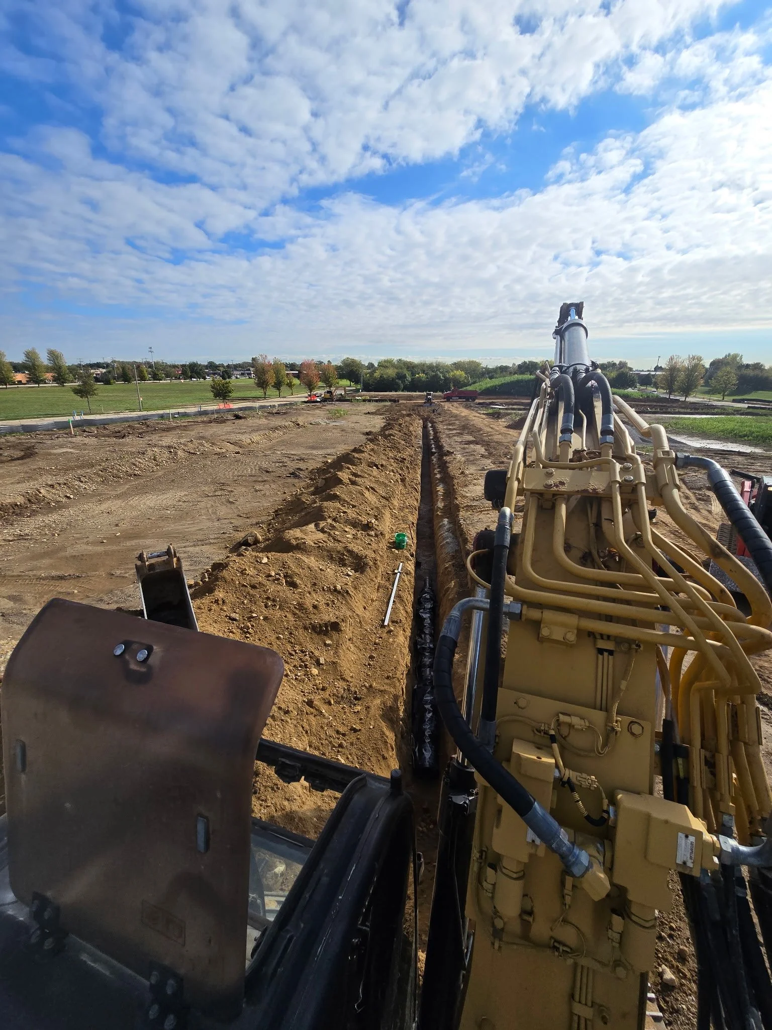 View from the operator's seat of construction equipment at a work site with dirt, a trench, trees, and a partly cloudy sky.