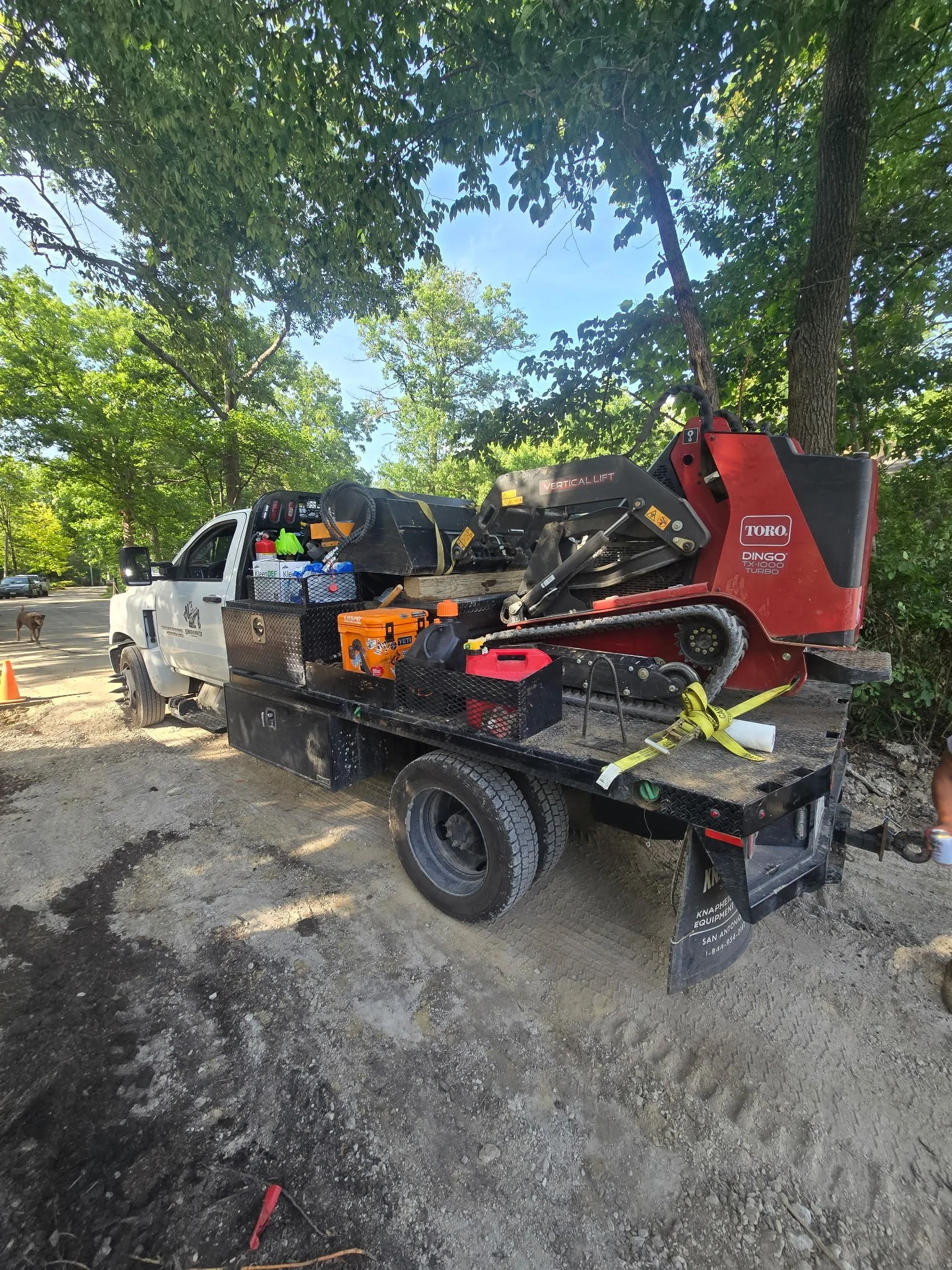 A work truck with construction equipment loaded on the back, parked on a dirt surface, with trees and blue sky in the background.