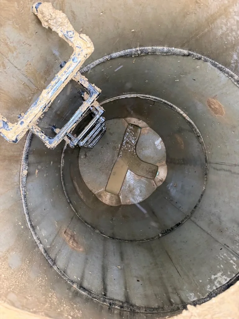 Looking down into an empty, cylindrical industrial tank or container with a metal paddle or agitator at the bottom, and a ladder or platform on the side.