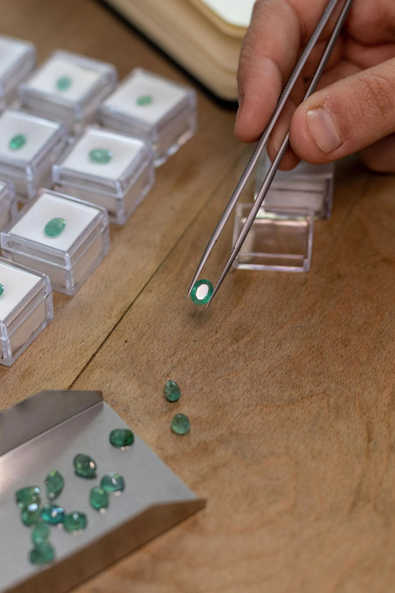 Close-up of a person's hand using tweezers to handle a small emerald gemstone, with several other emeralds on a metallic tray and a wooden surface nearby.