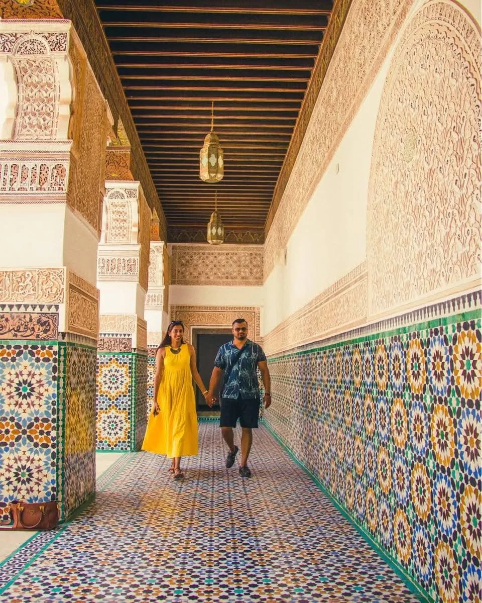 A couple holding hands walking through a corridor with colorful mosaic tile walls and a patterned floor in a building with intricate architectural details and hanging lanterns.