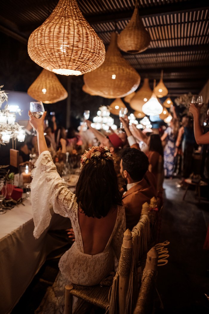 People raising glasses in a warmly lit, rustic wedding reception with woven pendant lights and floral decorations.