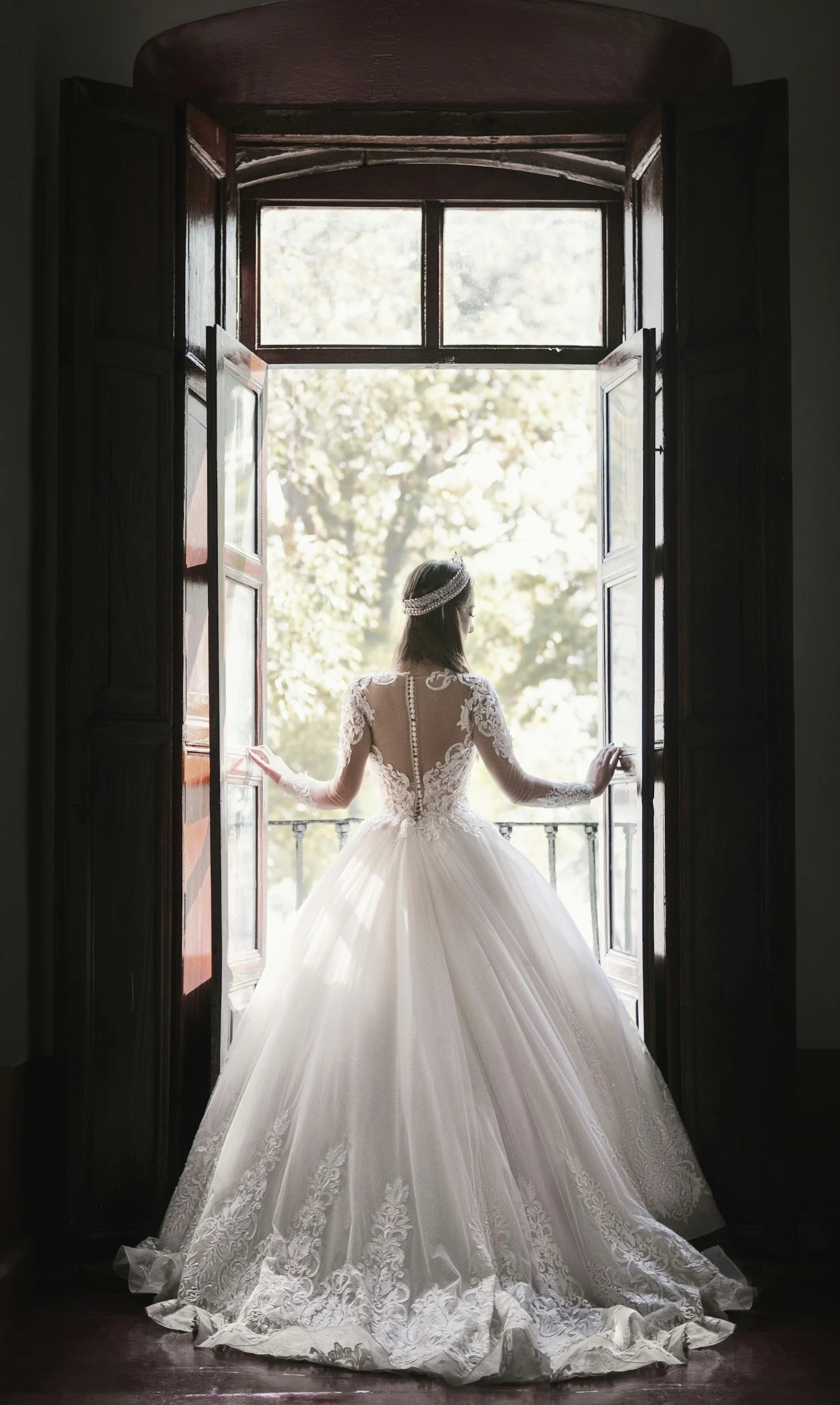 A bride in a white wedding gown standing in an open window, looking outside, with trees visible in the background.