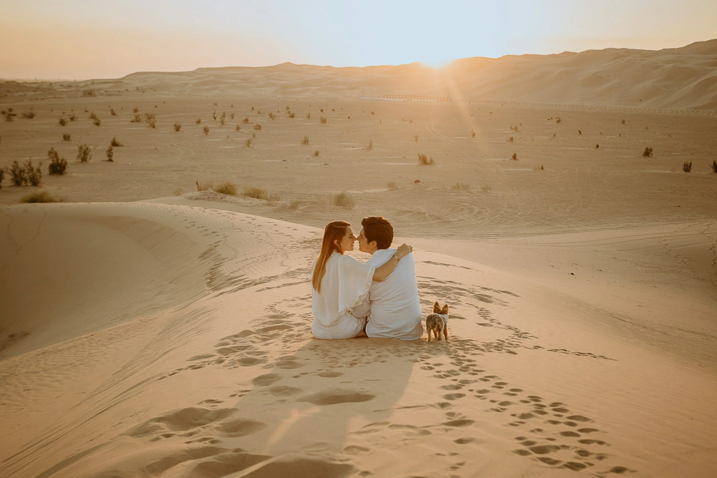 A couple sitting on sand dunes in a desert during sunset, embracing each other with a small dog nearby, and sparse desert vegetation in the distance.