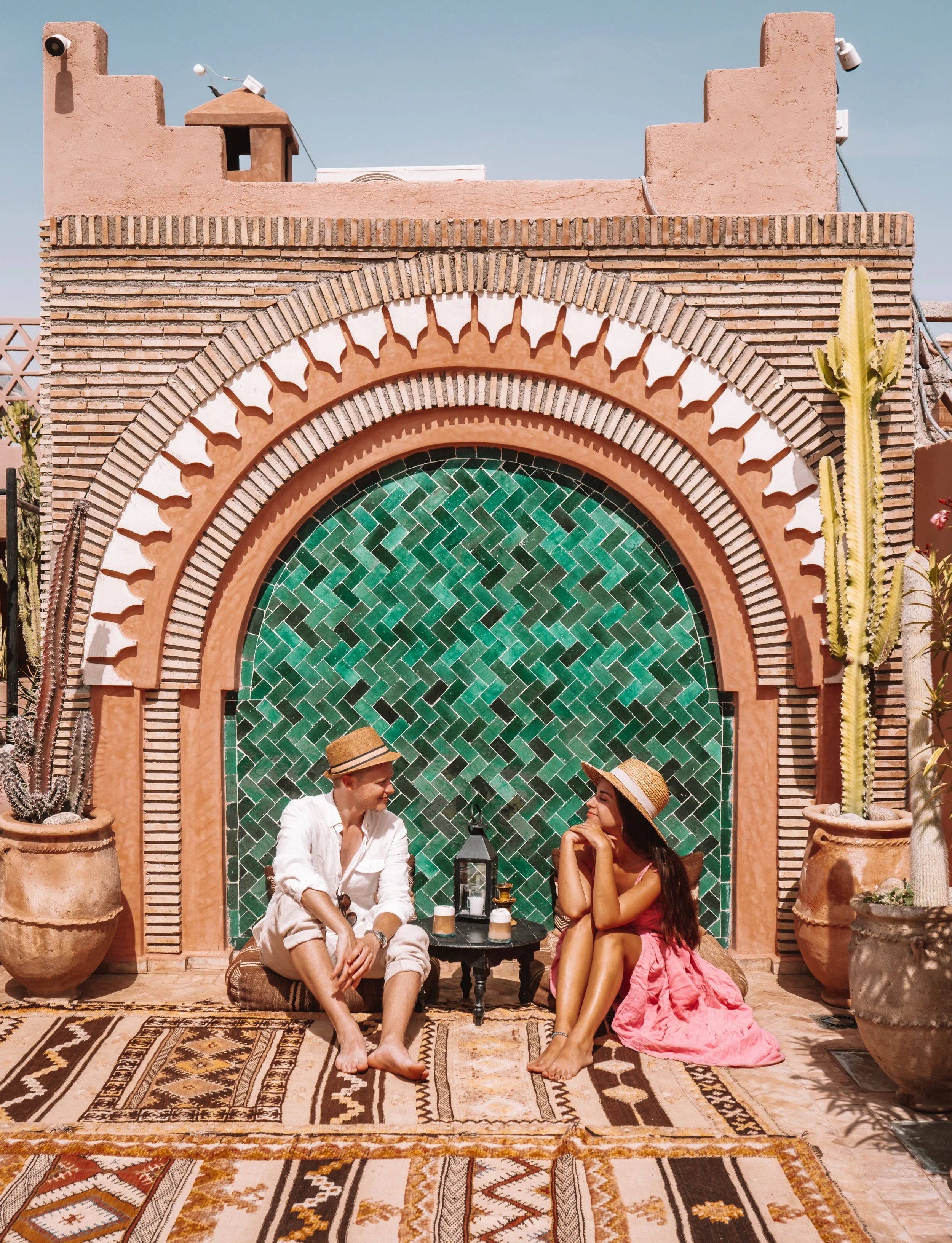 Two people sitting on a rug in front of a decorative arched wall with green tilework, large potted cacti on either side, and a small table with drinks. The man wears a white shirt and straw hat, the woman a pink skirt and straw hat, smiling and looking at each other.