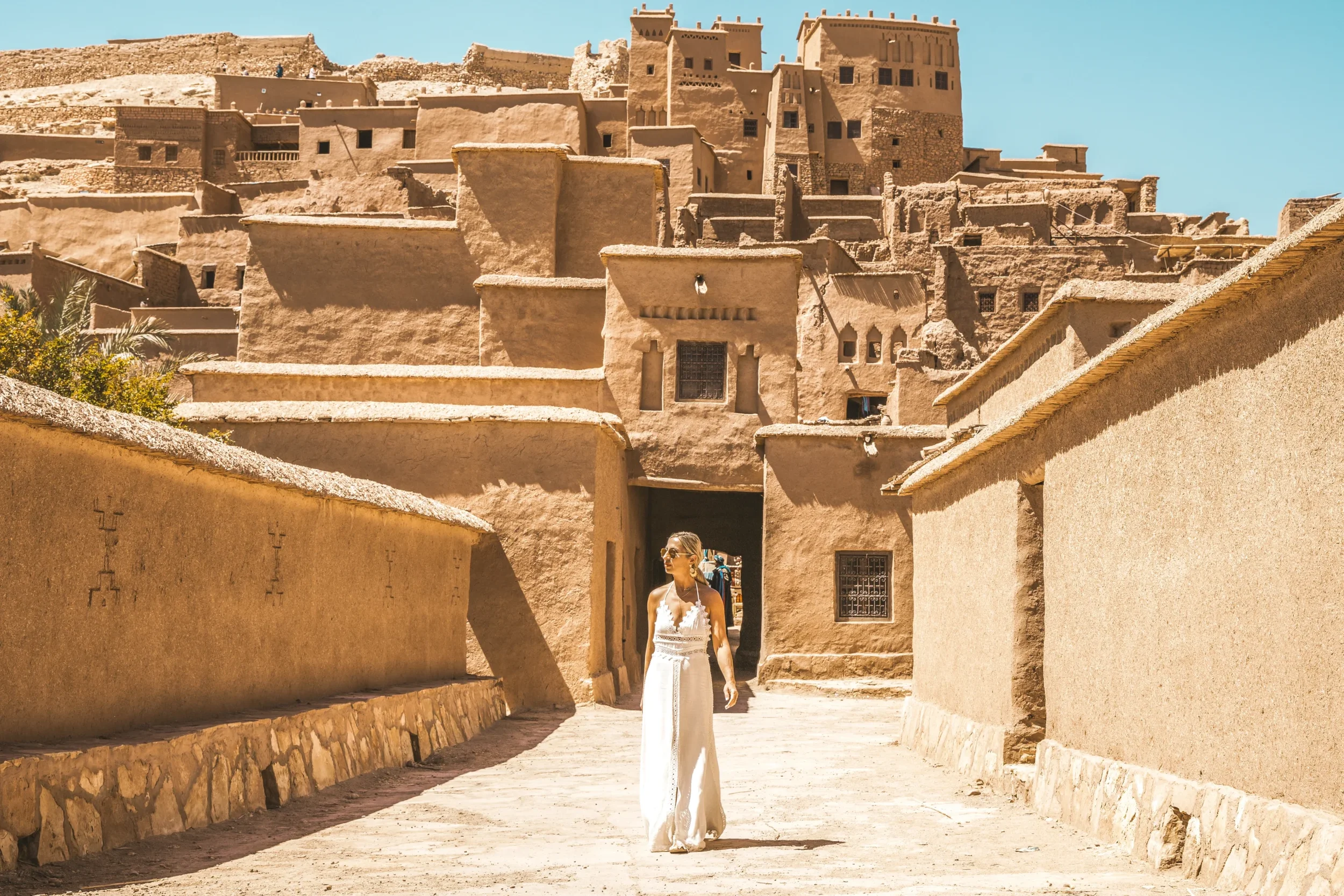 A woman in a white dress walking through an ancient desert town with adobe buildings and fortress walls under a clear blue sky.