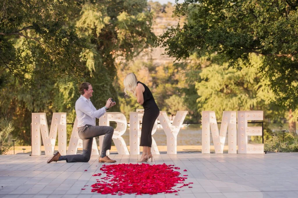Man proposing on one knee to his partner next to giant light-up MARRY ME letters and a red rose petal heart in Marrakech.