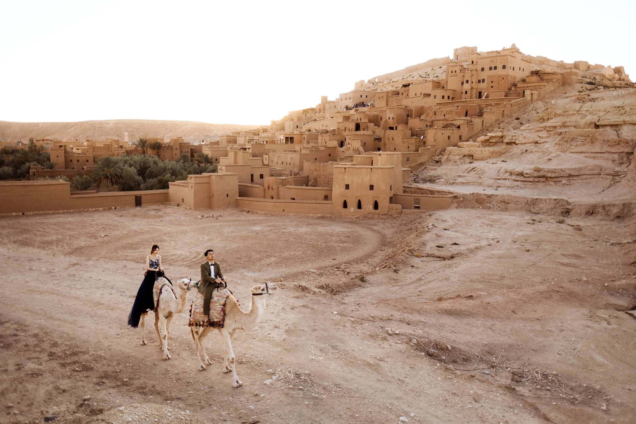 A desert cityscape with beige buildings built into hills. In the foreground, a woman and man ride camels across a dirt terrain. The woman is wearing a long dress, and the man is wearing a jacket with a bow tie. There is sparse vegetation and a few palm trees.