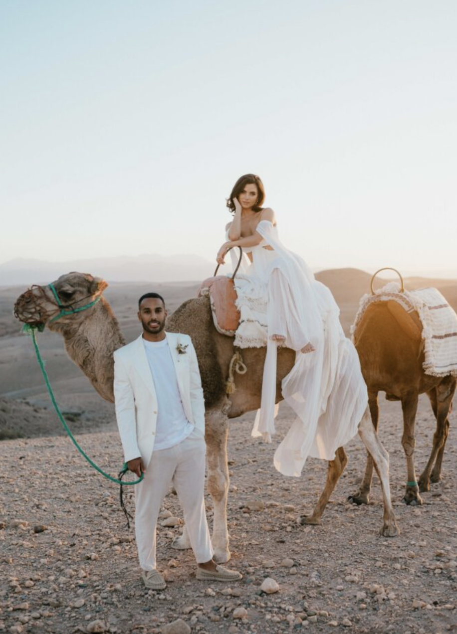 A couple in wedding attire with camels in a desert at sunset