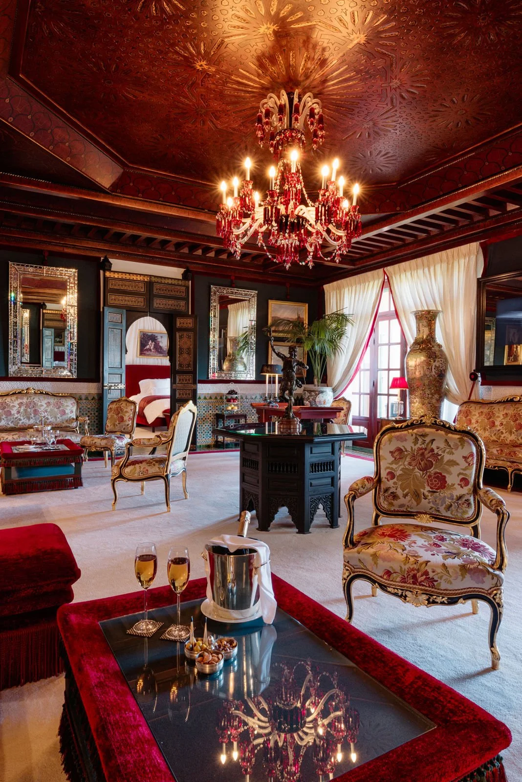 Elegant living room with floral upholstered chairs, a chandelier, large vase, draped curtains, and decorative mirrors.
