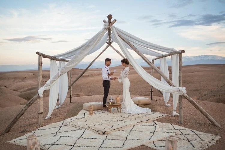 A couple getting married under a rustic driftwood canopy draped with white fabric, set in a desert landscape with distant mountains and cloudy sky.
