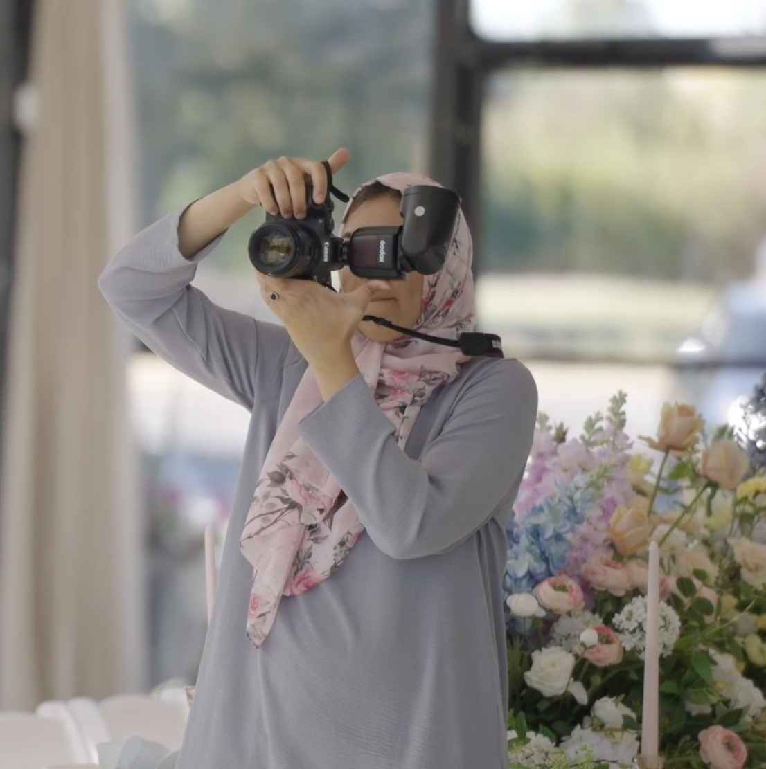 An elegant destination event photographer in a pink floral scarf holding a professional camera setup behind a luxury pastel floral centerpiece layout in Marrakech.