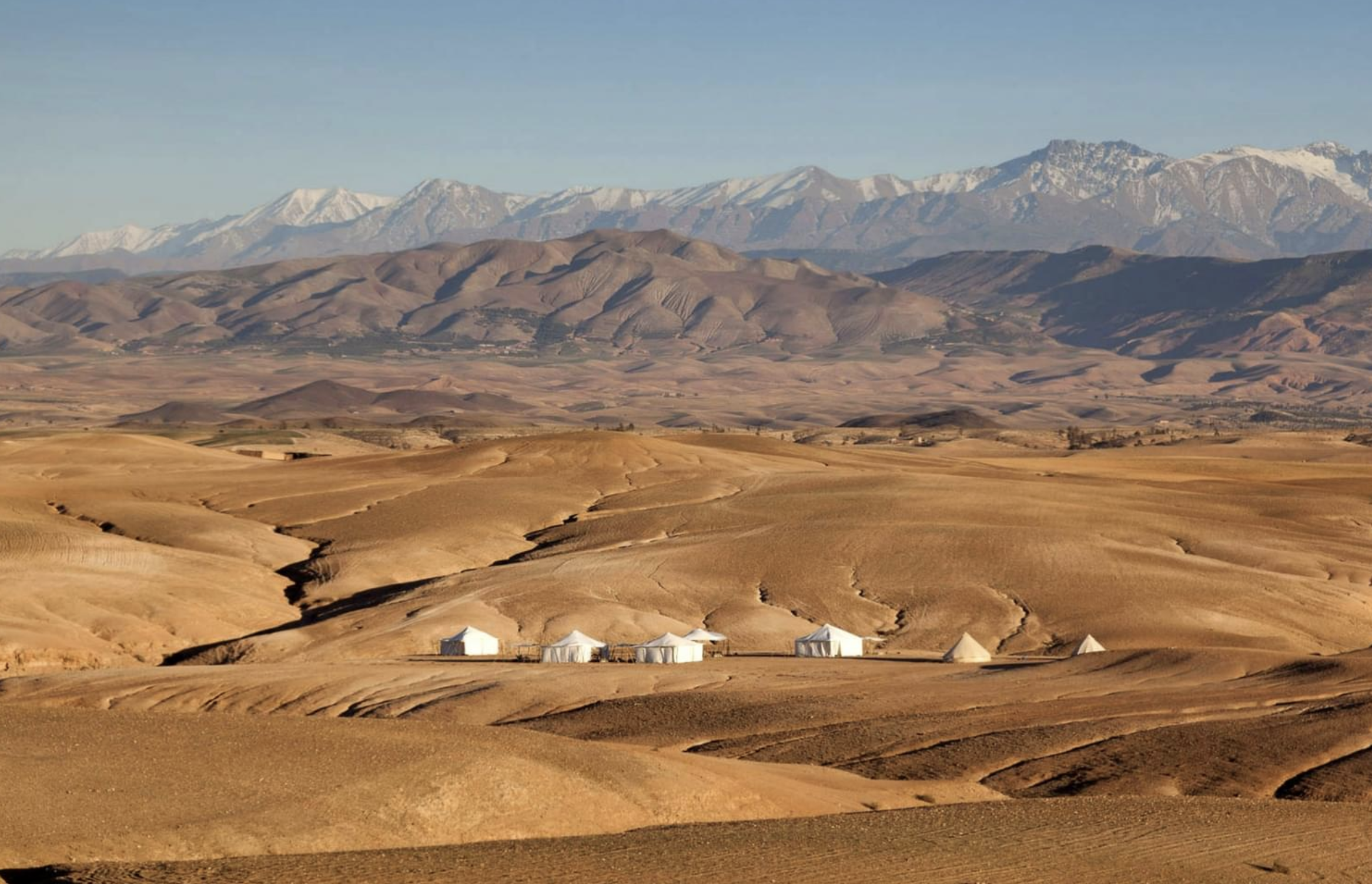 An expansive view of luxury white glamping tents nestled in the golden stone hills of the Agafay Desert with the snow-capped Atlas Mountains in the background.