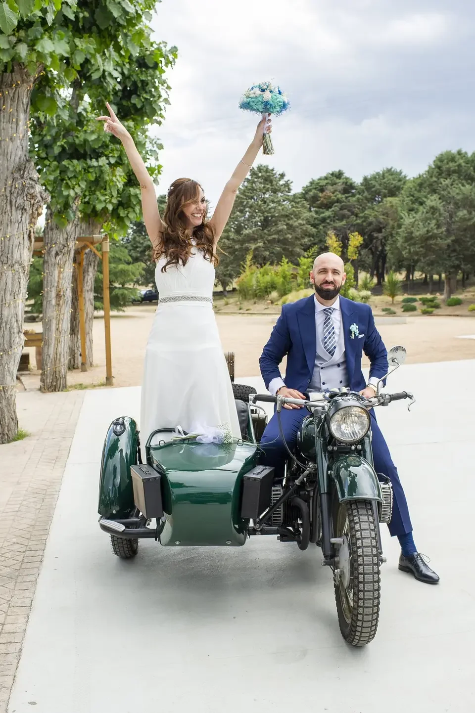 A bride and groom on a vintage motorcycle with a sidecar outdoors on a cloudy day. The bride is standing in the sidecar, raising a bouquet in the air, smiling, and the groom is sitting on the motorcycle, dressed in a suit, looking at the camera.