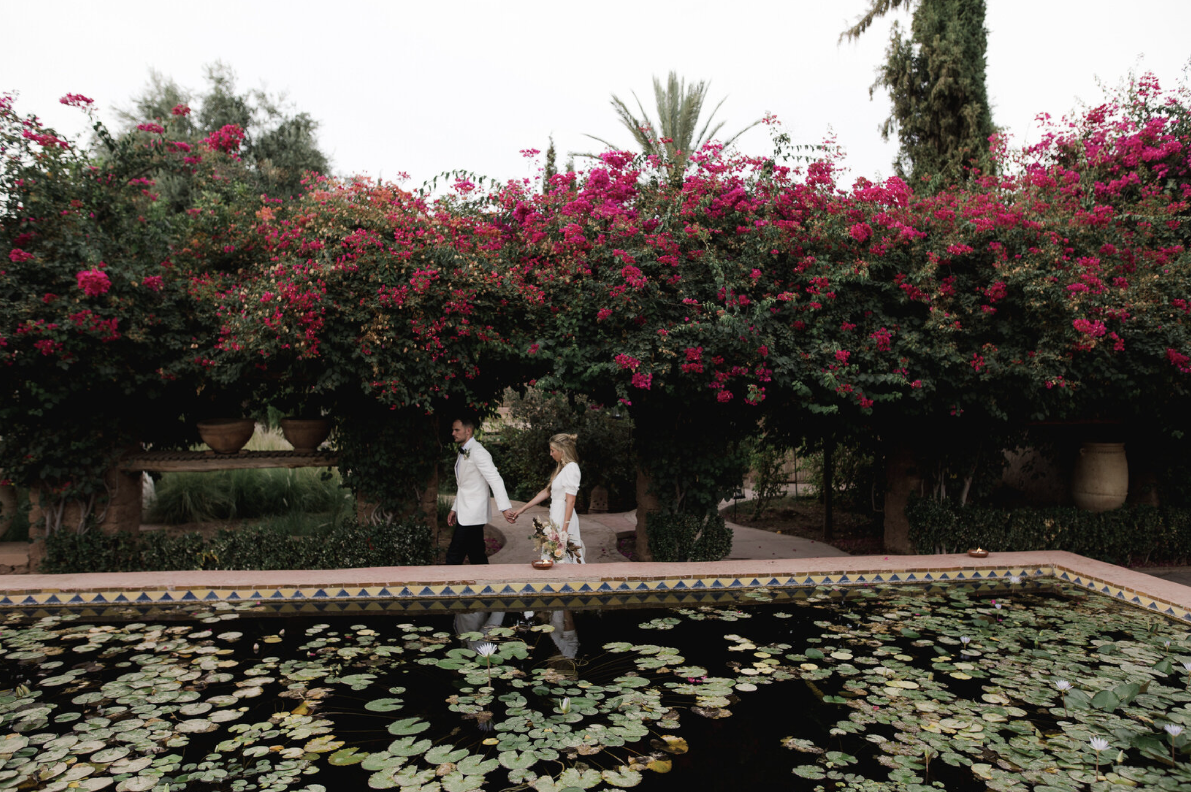 A couple dressed in white wedding attire holding hands and walking near a pond with water lilies, beneath large pink flowering bushes and surrounded by greenery.