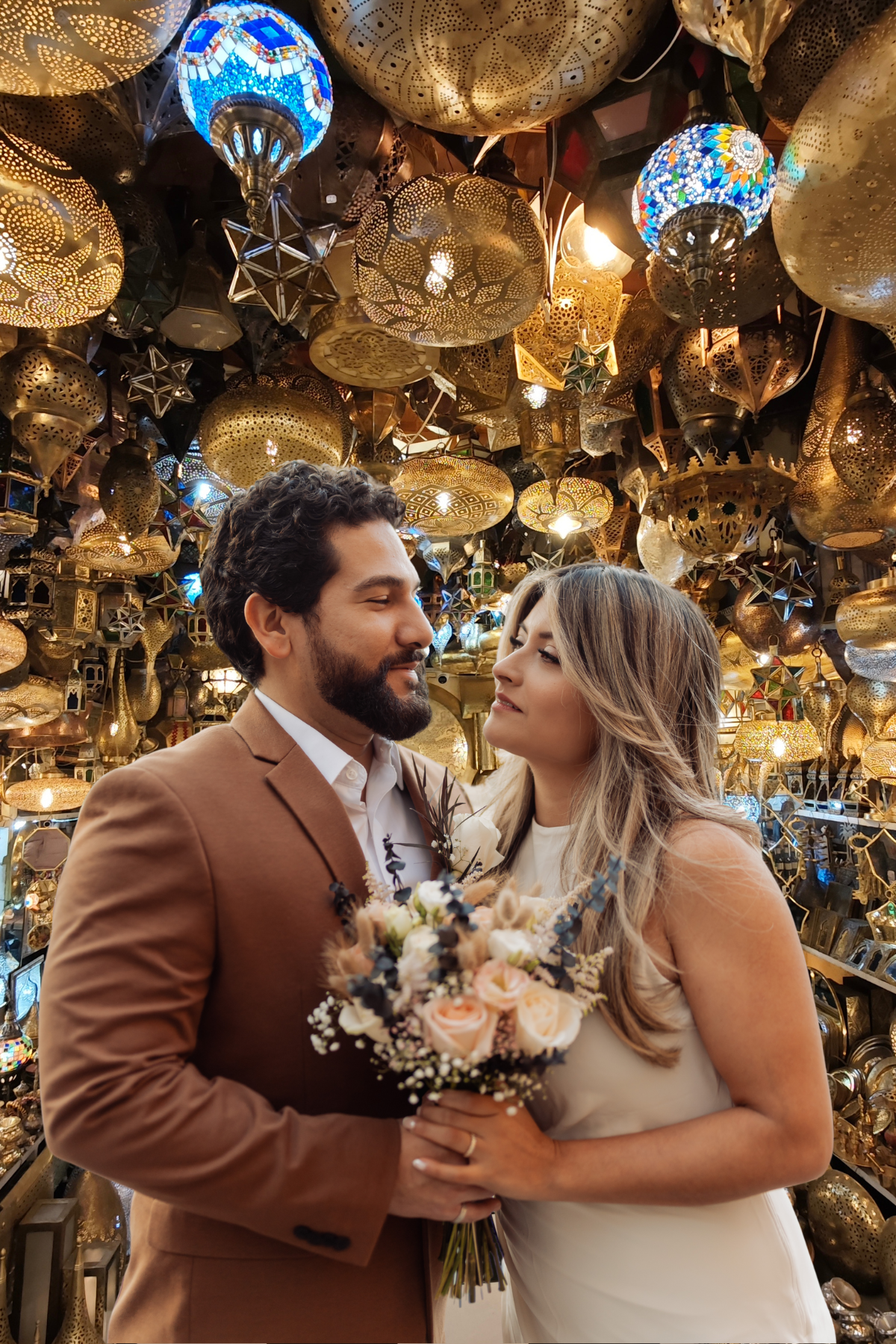 A couple dressed in wedding attire standing close together, holding a bouquet of flowers, with intricate decorative hanging lamps in shades of gold and blue overhead.