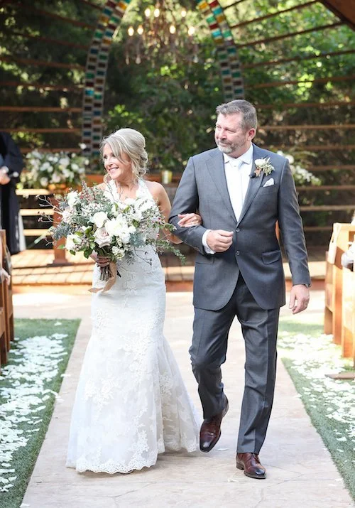 A bride wearing a white lace wedding gown holding a large bouquet walking down the aisle with a groom in a gray suit, at an outdoor wedding ceremony with a wooden arch and green trees in the background.