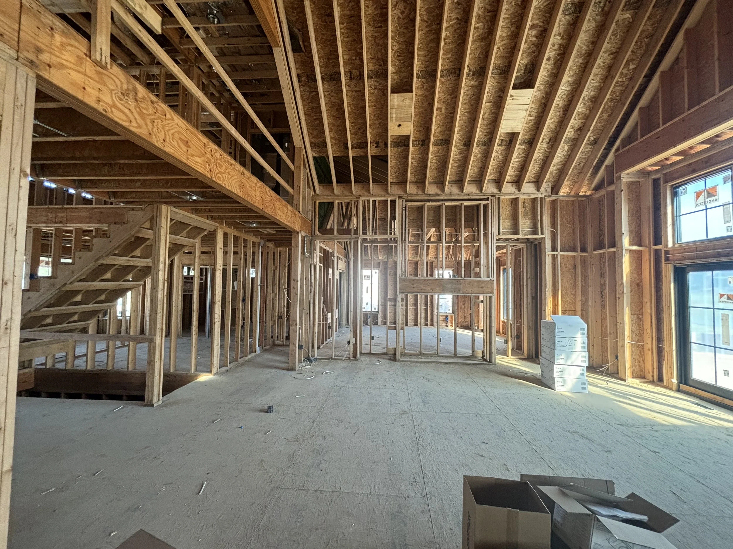 An interior view of a custom residential project by Booras Builders showcases active rough-in framing, featuring a vaulted ceiling with exposed wooden rafters, a framed staircase structure, and open stud walls over plywood subflooring.