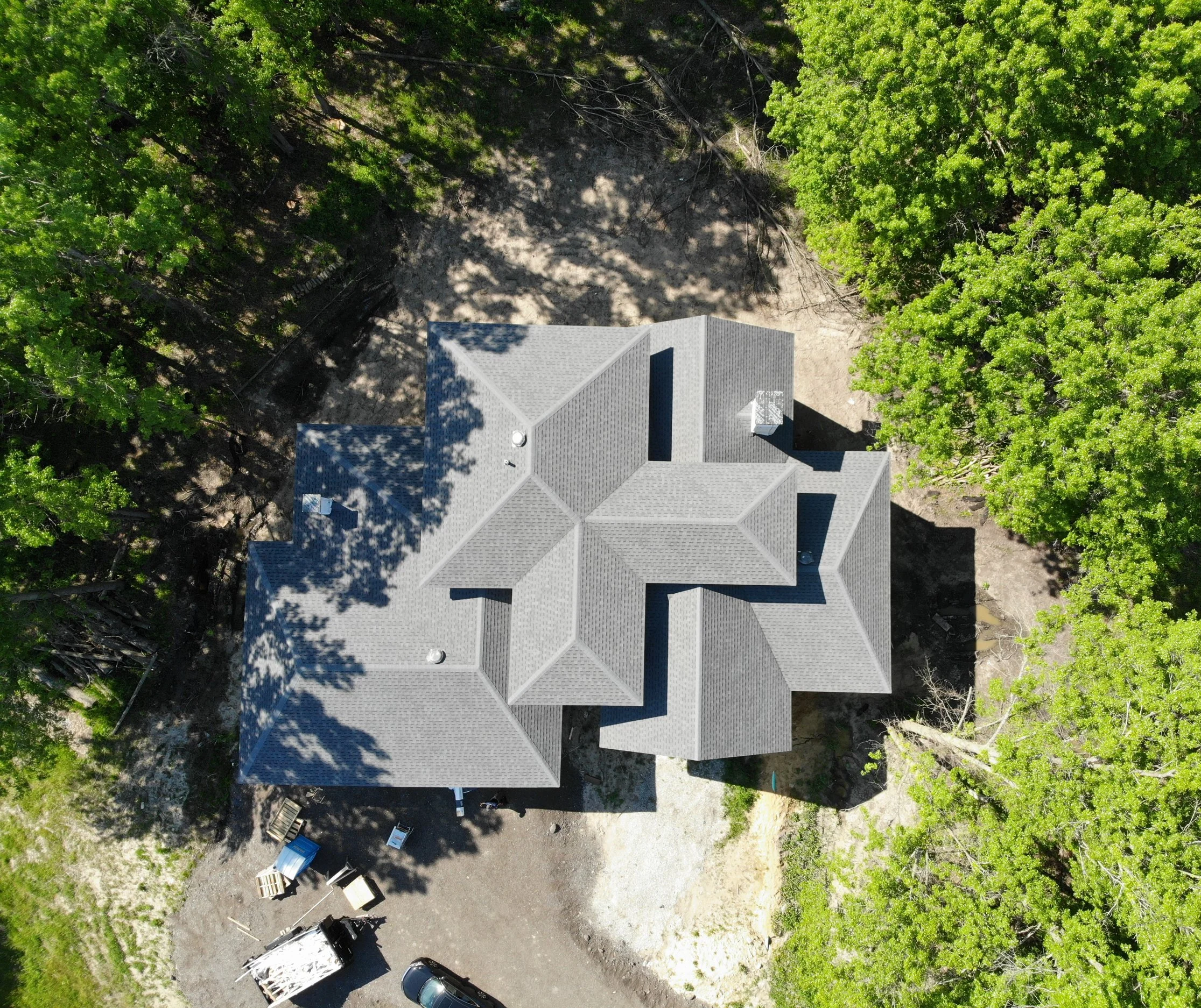 Surrounded by dense, mature trees, a freshly installed architectural shingle roof showcasing a complex multi-gabled design anchors this custom residential build that was both designed and completed by Booras Builders.