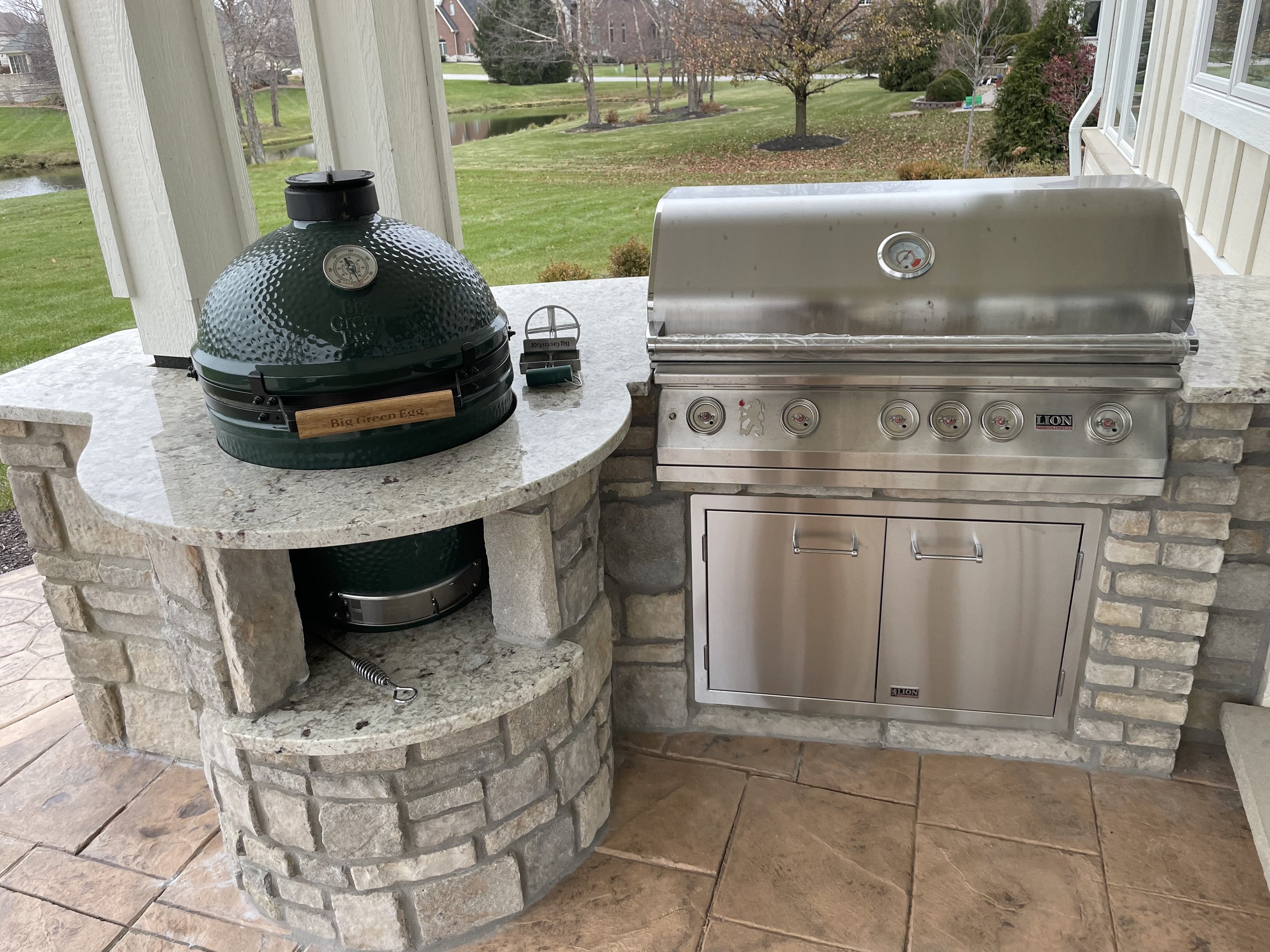 This custom outdoor barbecue station by Booras Builders includes a stainless steel grill and a smoker into a curved stone island with a polished granite countertop, set on a stamped concrete patio.