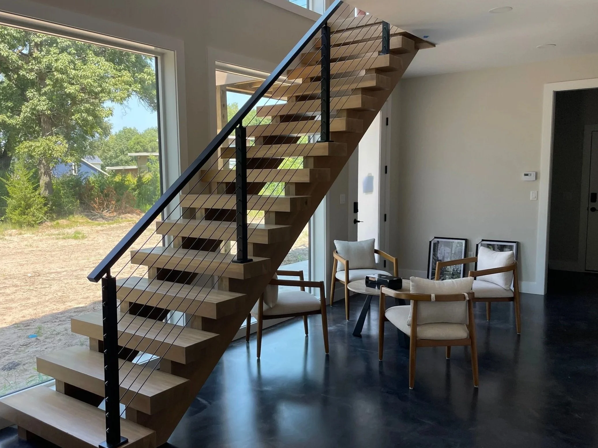 Interior of a modern home in Porter County featuring an architectural floating staircase with thick wooden treads and black metal handrails and cables, set against dark polished flooring and an expansive picture window.