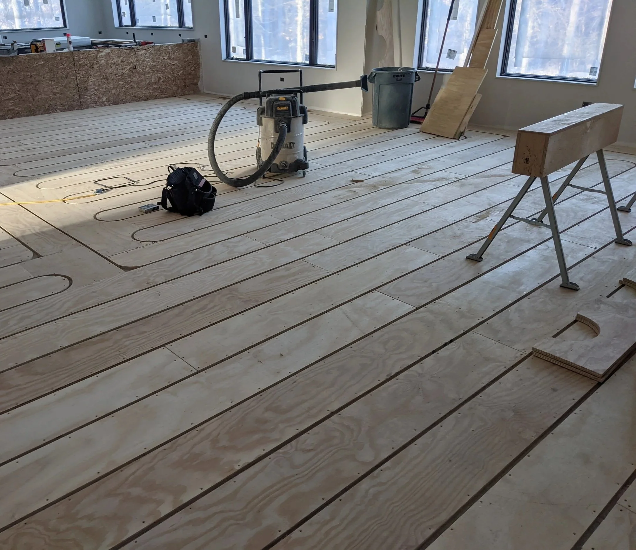 A family room under construction with wooden flooring, a vacuum, trash bin, and wooden boards. There are thin gaps in the flooring where tubes will be placed to warm the home by radiant heat, a luxury custom-built into this Booras Builders home.