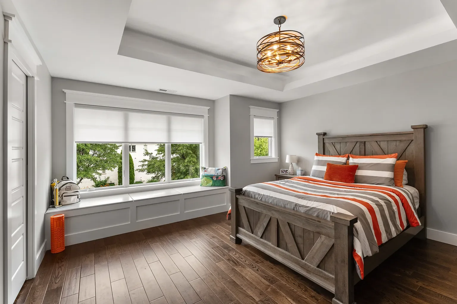 This bedroom, part of a residential project in Chesterton by Booras Builders, spotlights a tray ceiling with an orb light fixture, a built-in window seat alcove with wainscoting paneling, and clean white trim contrasting against the hardwood flooring