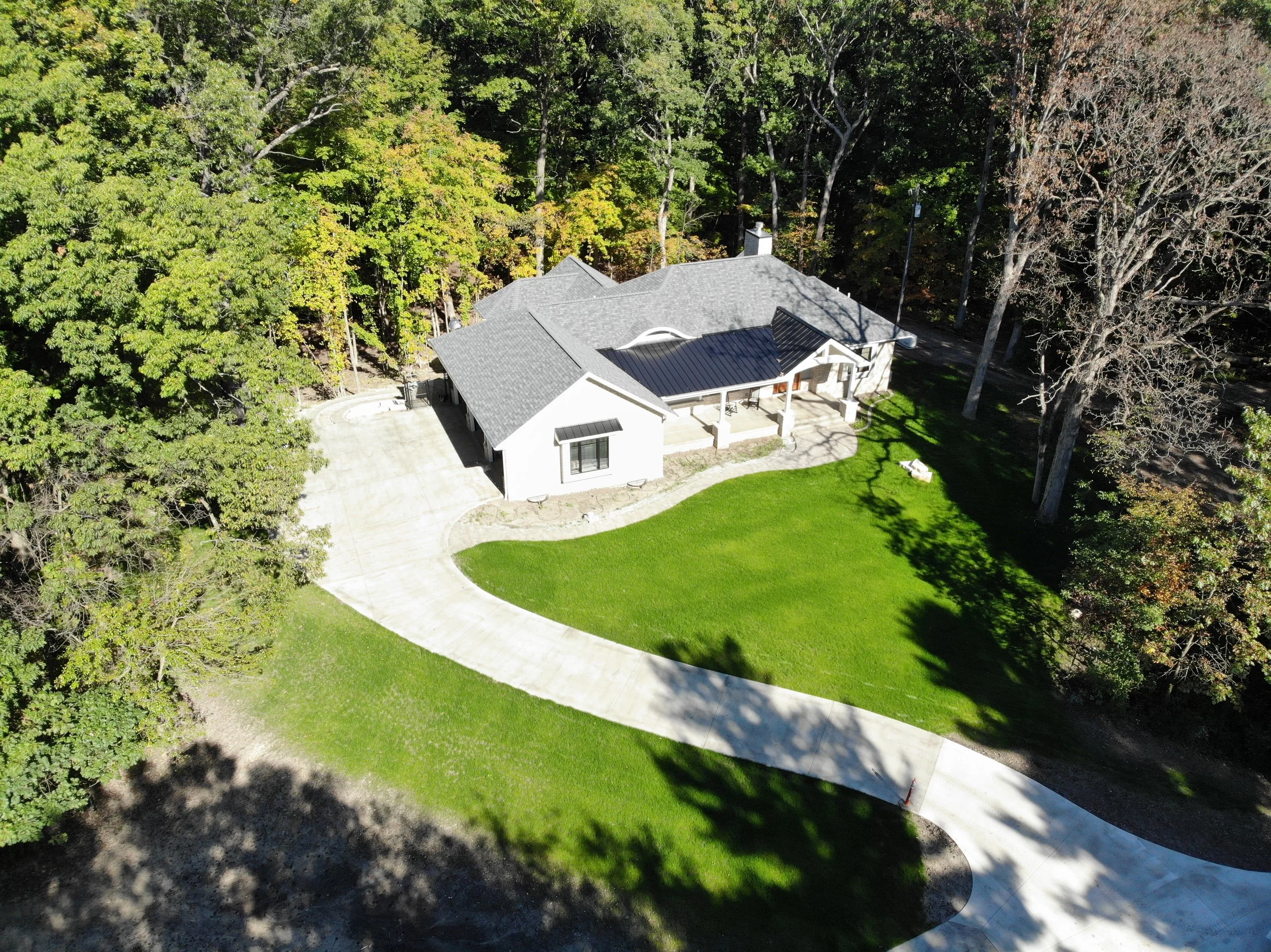 An aerial view of a house surrounded by trees and green lawn, with a concrete driveway leading to the front entrance.