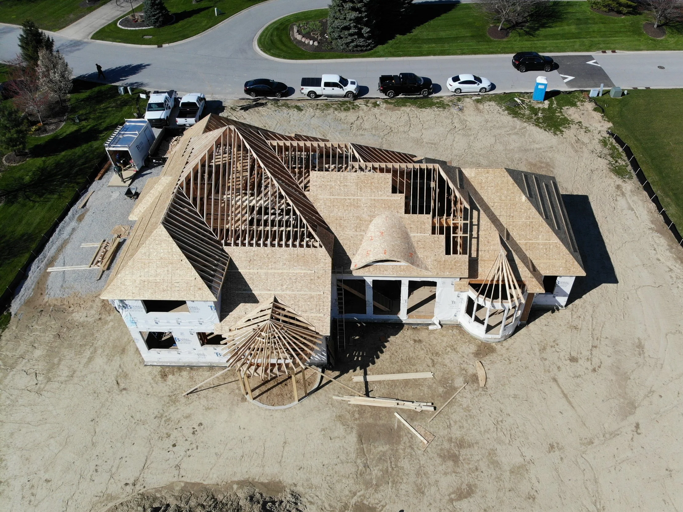 An aerial drone view captures the detailed roof framing layout of a custom estate under construction by Booras Builders in Northwest Indiana, highlighting roof valley intersections, a complex eyebrow dormer, and multiple circular turret structures.