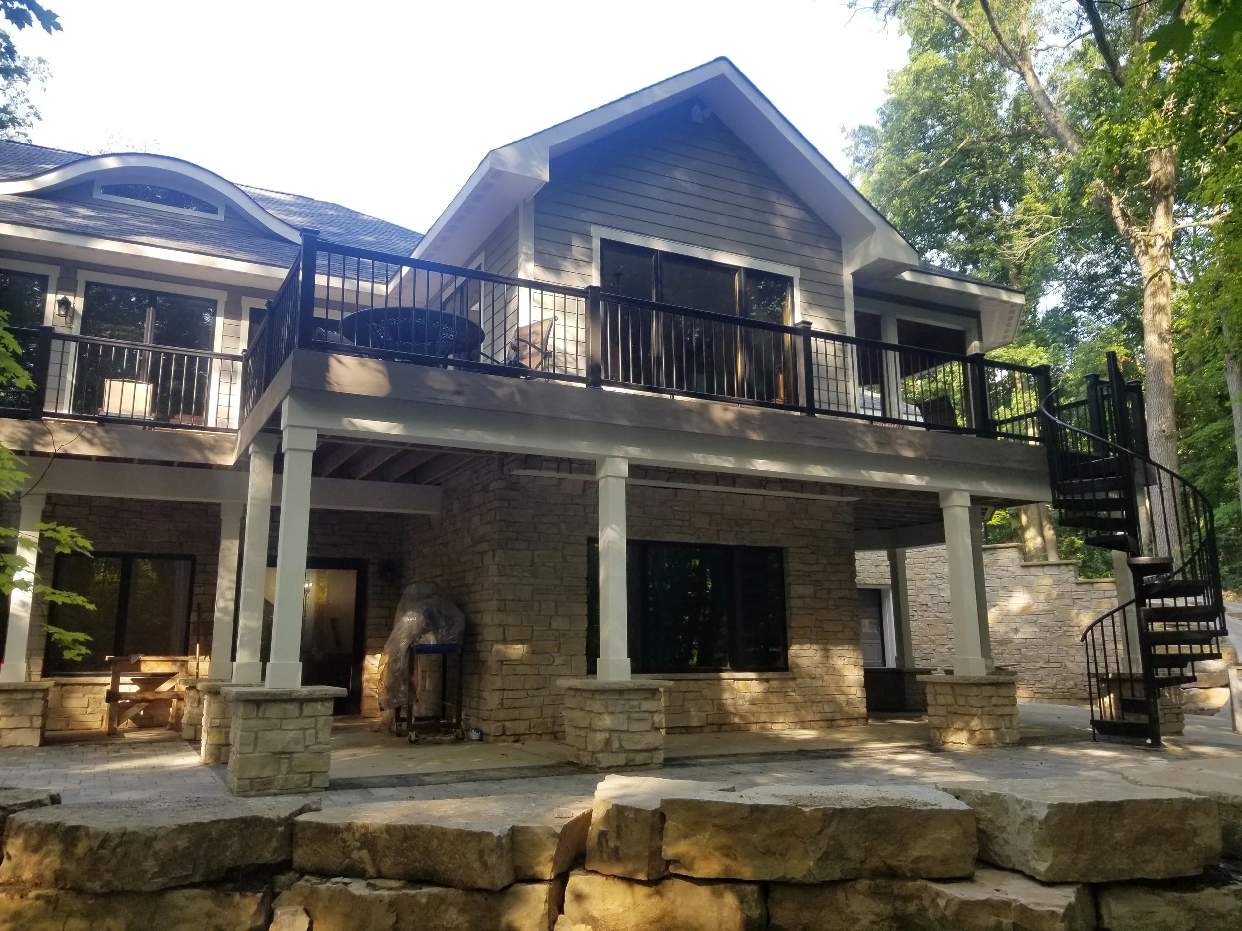 In Porter County, Booras Builders built a custom-built home whose rear exterior showcased a spacious second-story deck with black railings, a black spiral staircase, a covered lower-level patio, stone masonry, and a wooded landscape.