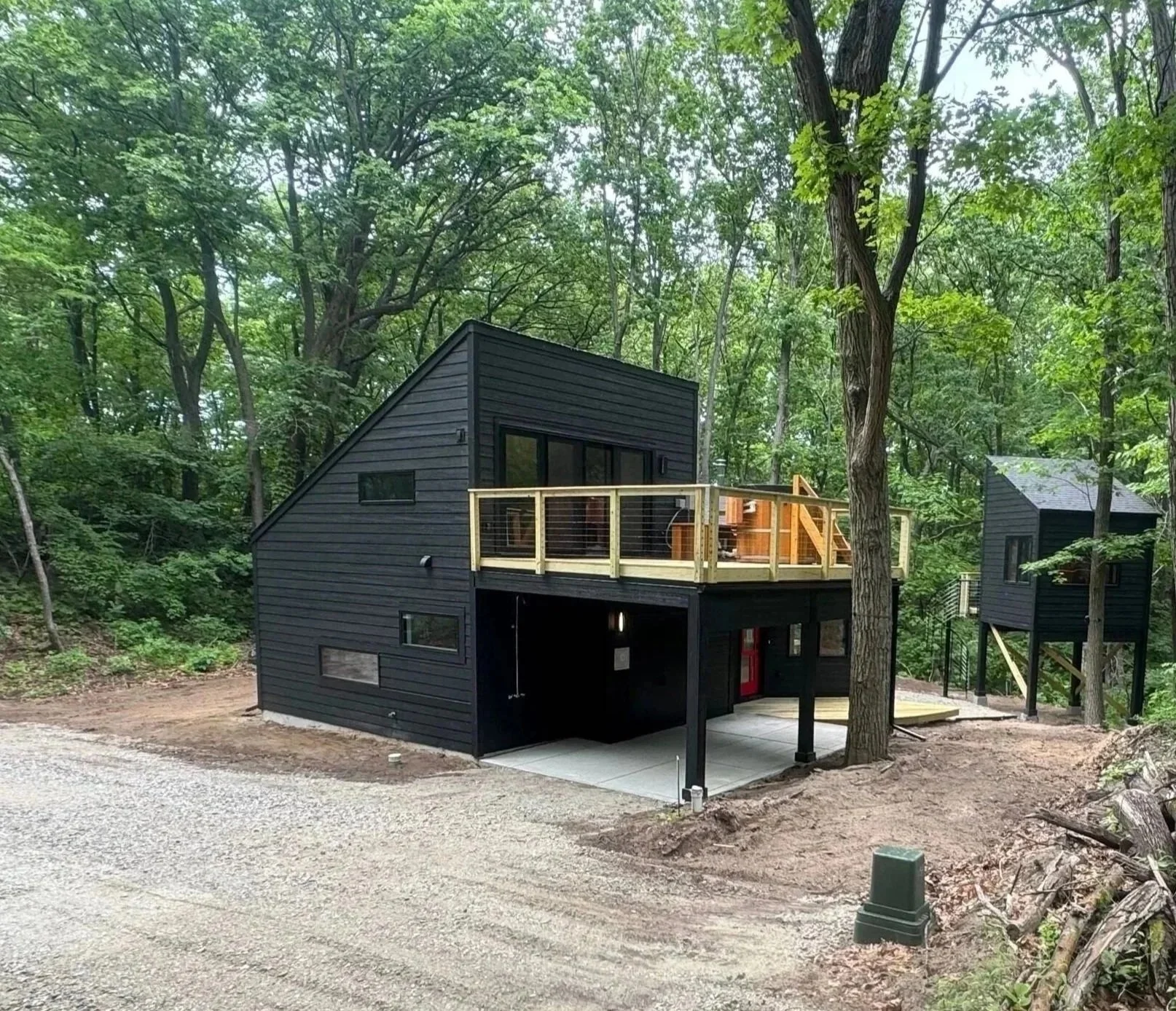 This sleek, modern dark cabin has a slanted roof and large windows. It's located in a forest in Gary, Indiana and is energy efficient. Elevated deck above an open carport area. A pile of cut logs sits on gravel to the right.